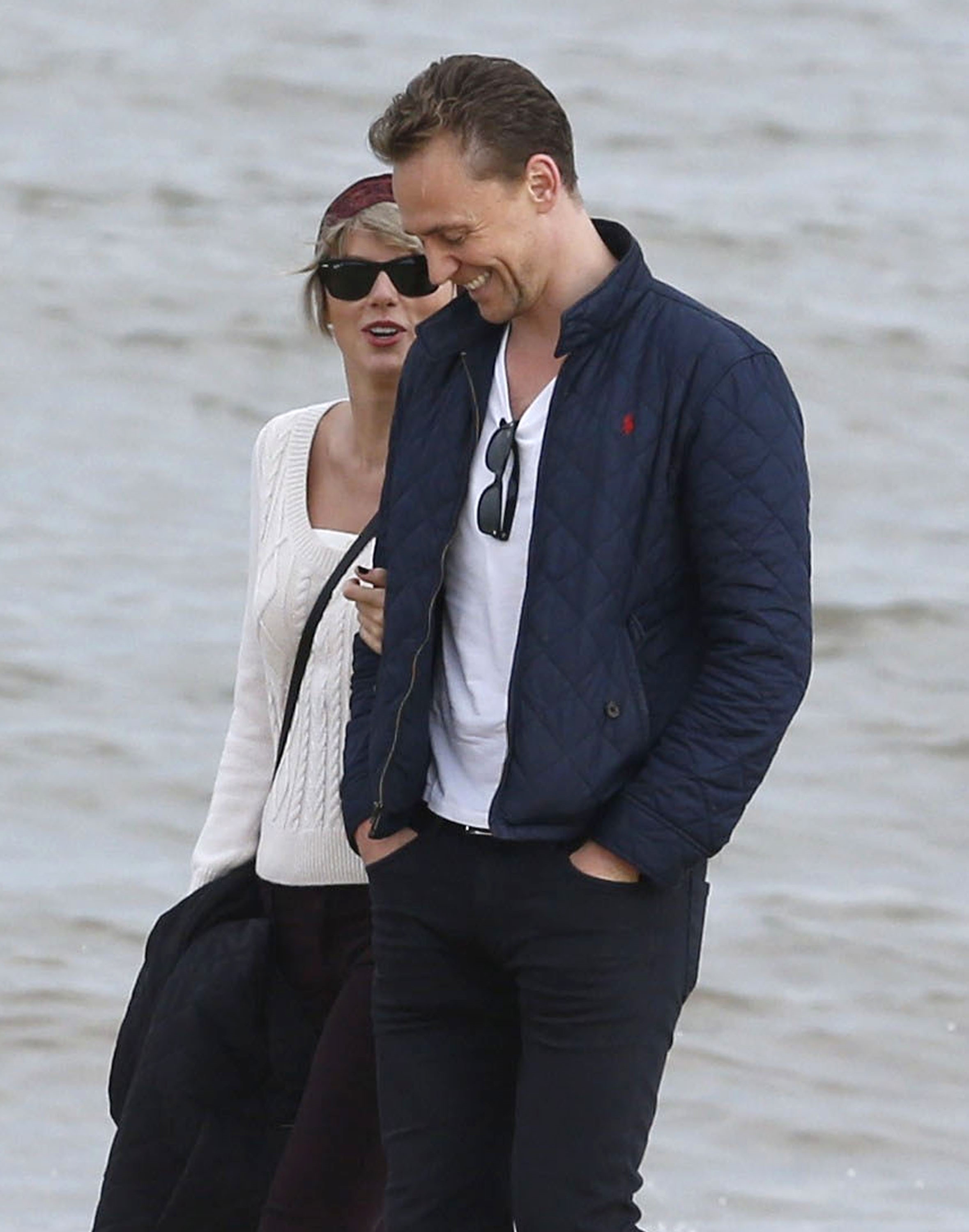 Taylor Swift and Tom Hiddleston walk and smile together on a beach, dressed casually. Taylor wears a knit sweater and sunglasses, while Tom has a jacket and sunglasses