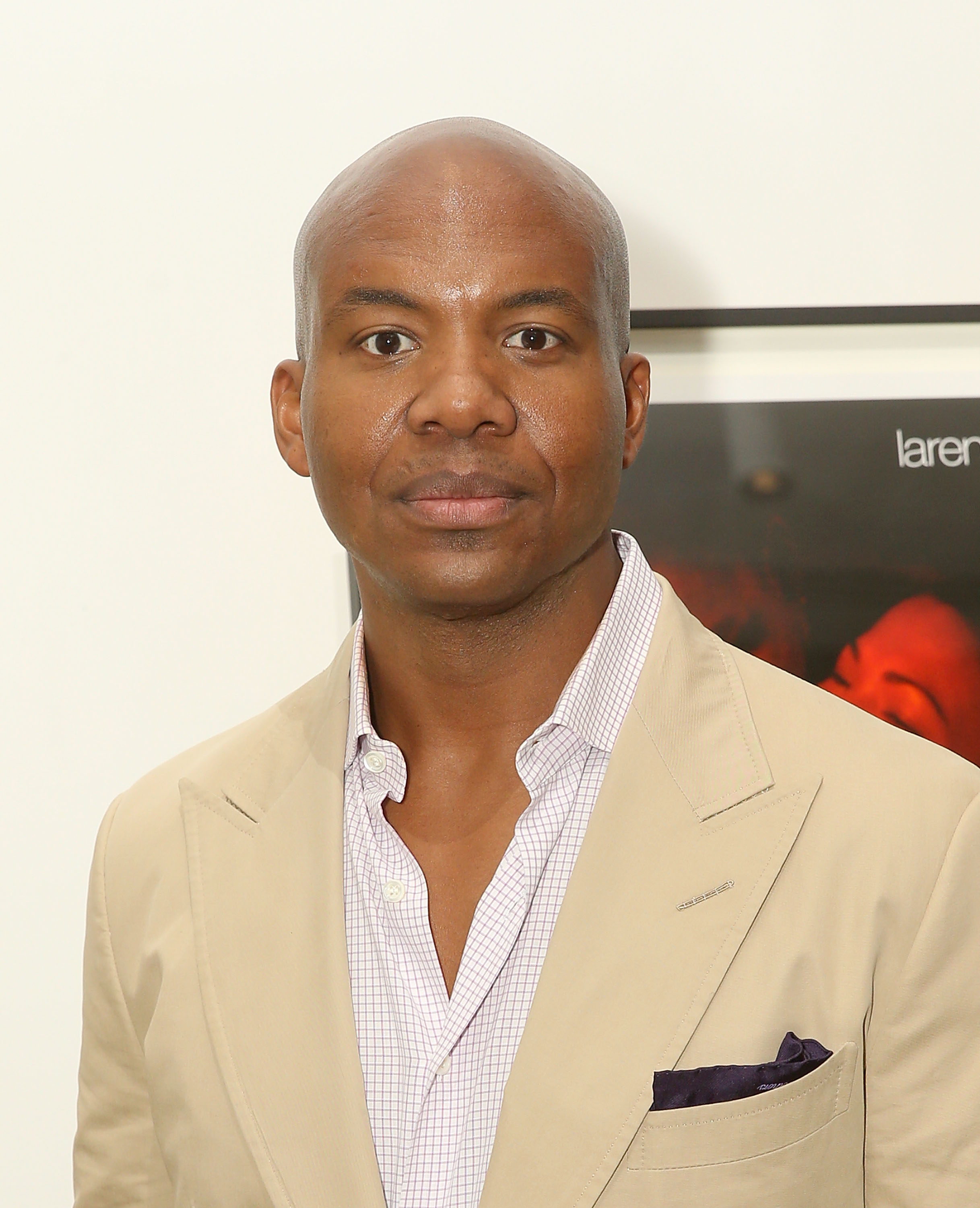 A man in a beige suit with a light checkered shirt, standing indoors in front of a framed picture