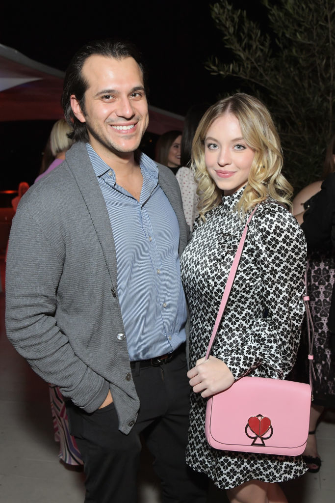 Jonathan Davino in a blazer stands next to Sydney Sweeney, who is wearing a patterned dress and carrying a purse with a heart design
