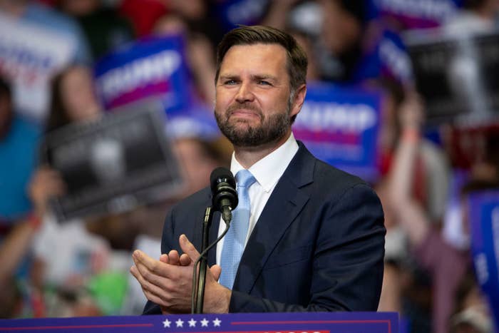 J.D. Vance stands at a podium, clapping with a slight smile, while supporters holding campaign signs are visible in the background