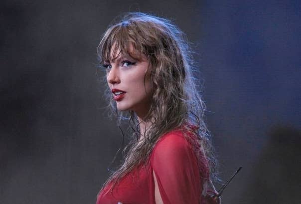 Taylor Swift on stage wearing a stylish outfit with flowing sleeves, reaching out to the audience during a performance. The background is misty