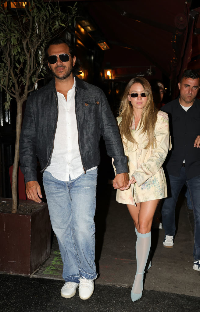 Jonathan and Sydney walk hand-in-hand at a nighttime event, with him wearing a casual jacket and jeans, and her in a stylish, short coat and thigh-high boots