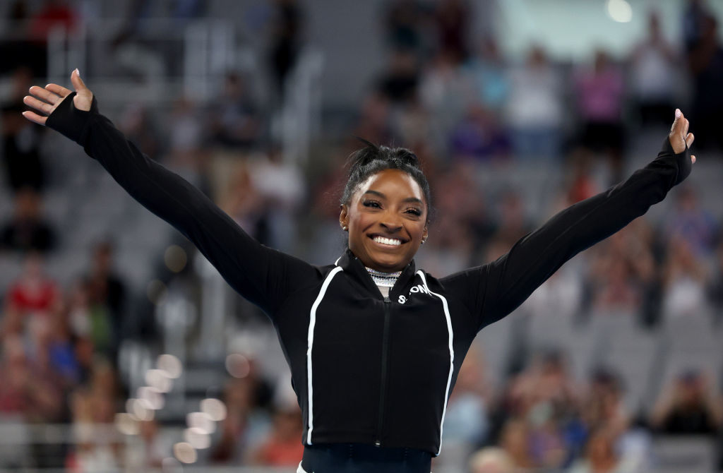 Simone Biles with arms raised, smiling, wearing an athletic jacket at a sporting event crowd in the background