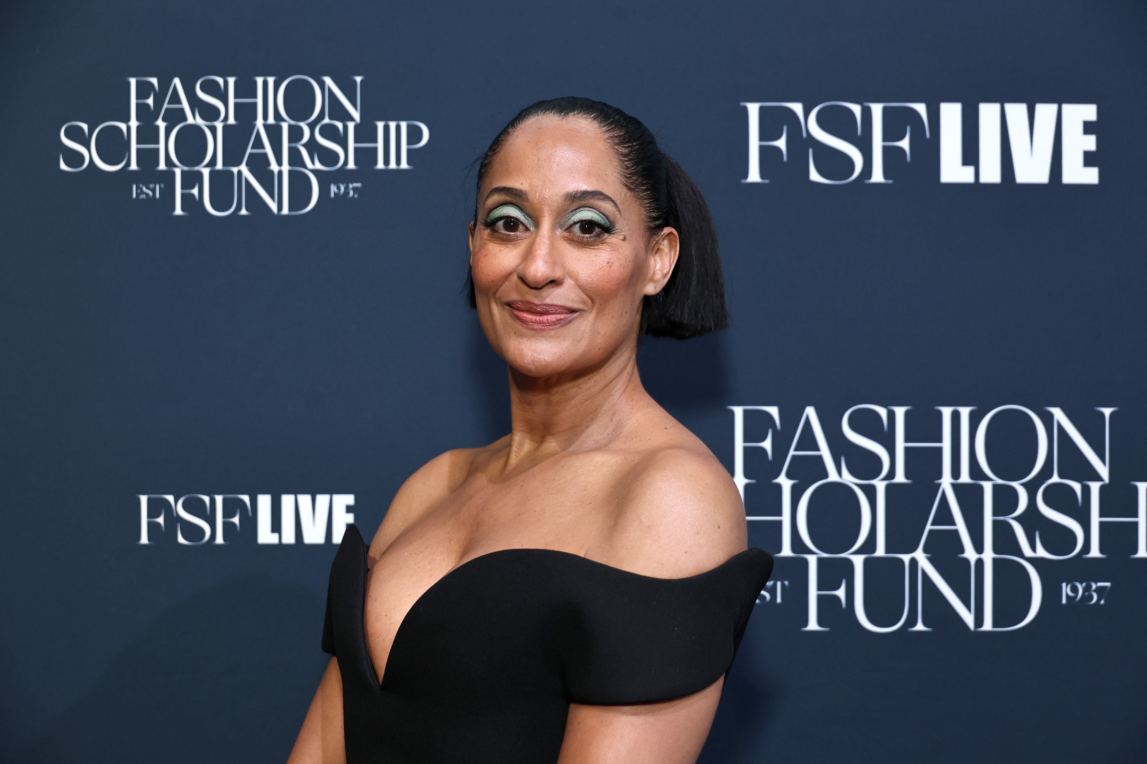 Tracee Ellis Ross poses in an off-shoulder black dress at the FSF Live event, with Fashion Scholarship Fund signage in the background
