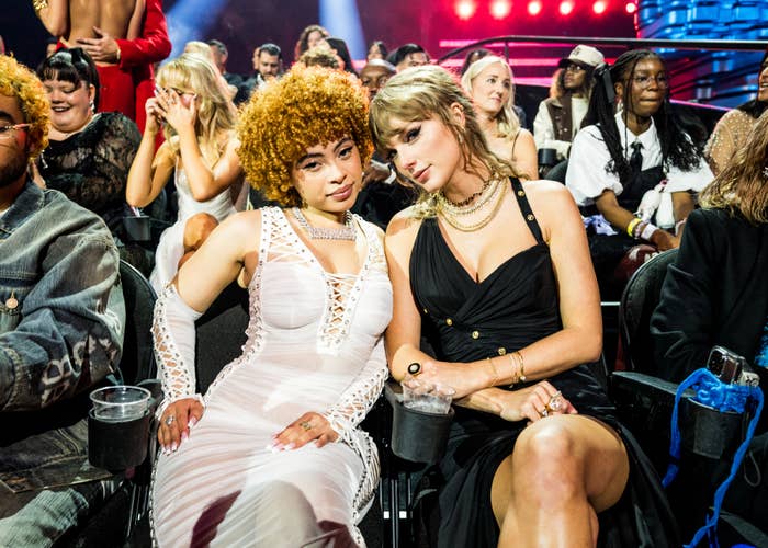 Ice Spice in a lace-up dress and Taylor Swift in a dark dress with gold accessories sitting close together in an event audience