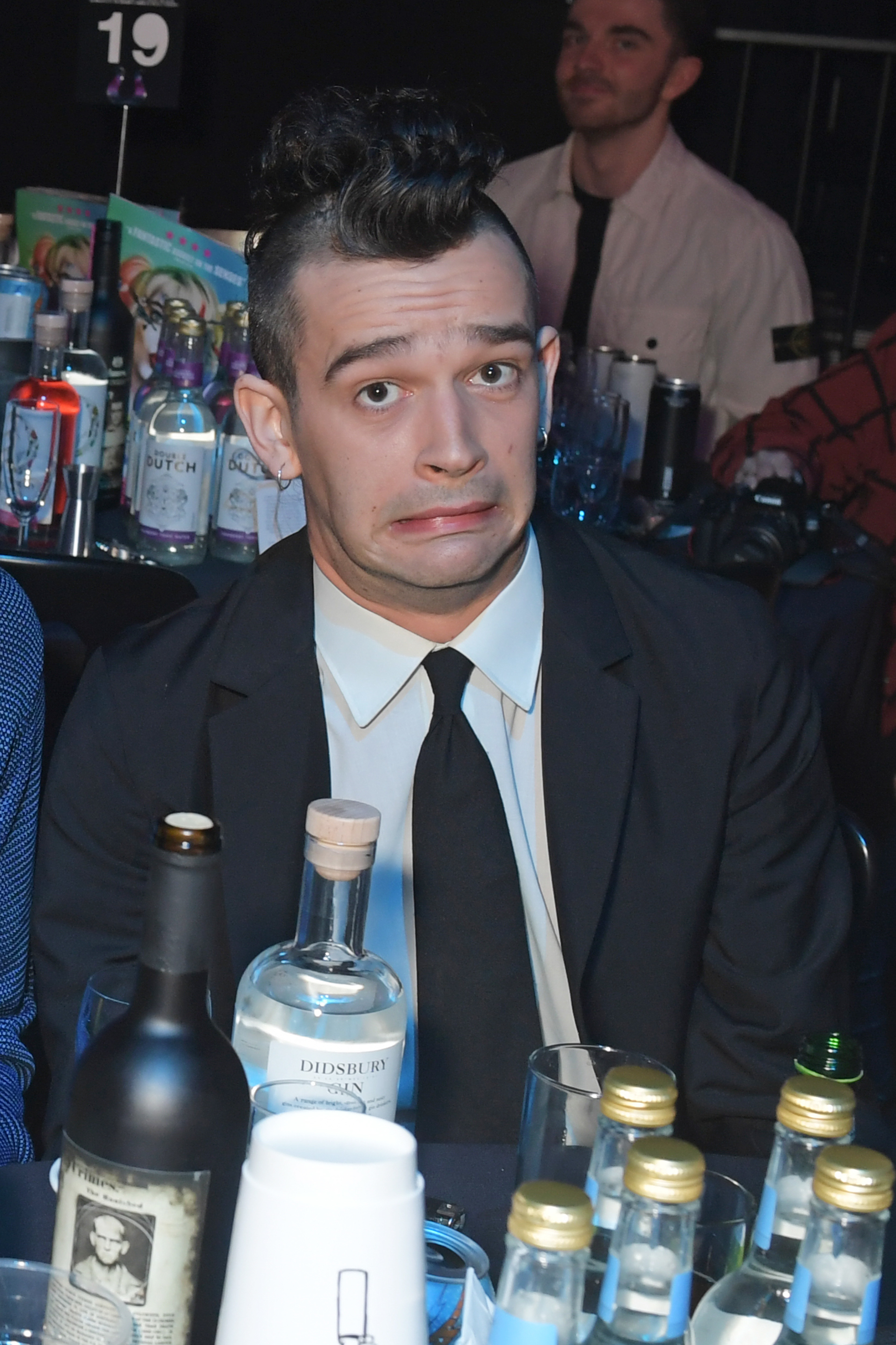 Matty Healy sits at a table making a funny face, wearing a suit and tie, with various drinks and bottles around him at an event