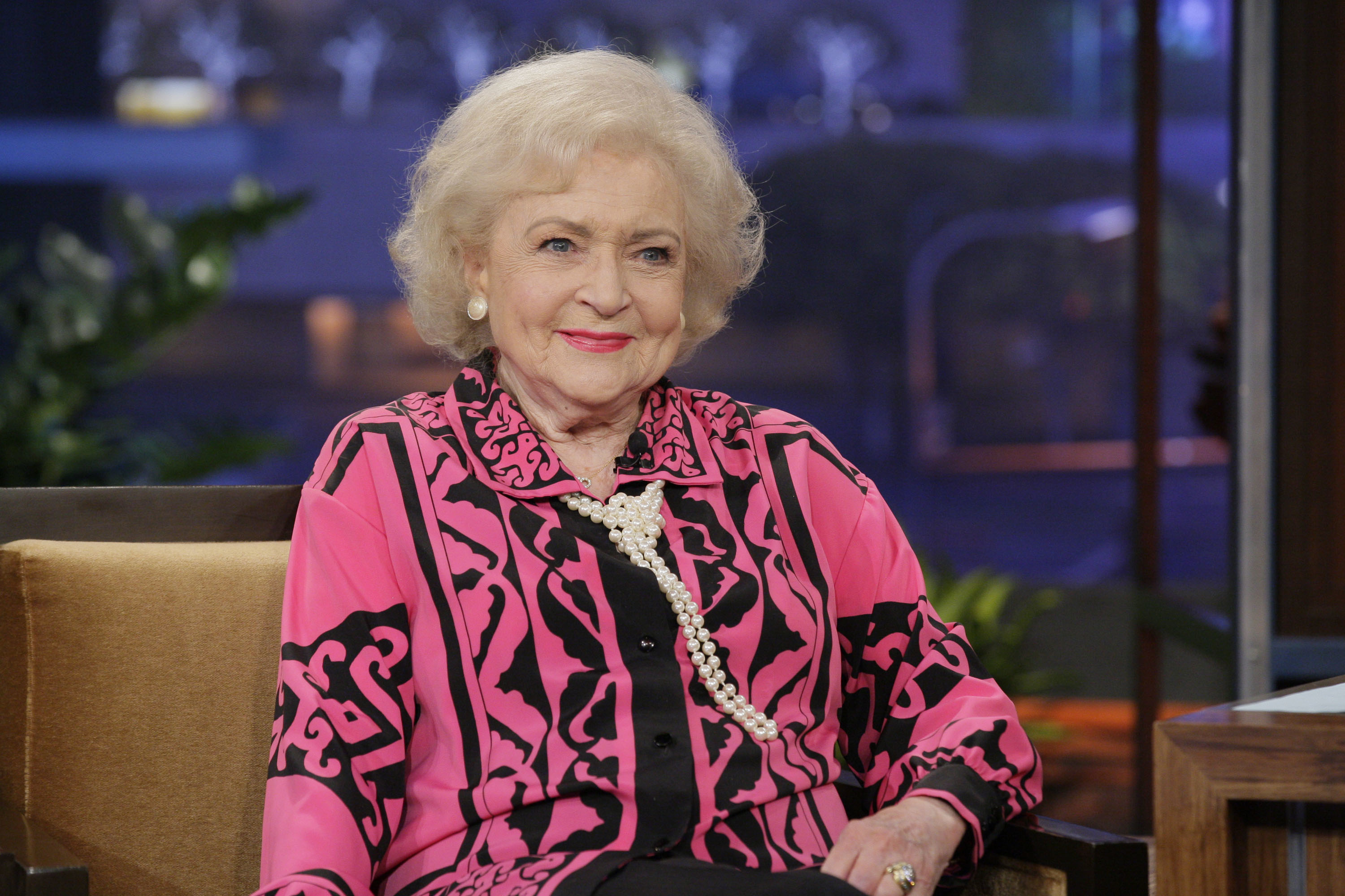 Betty White sitting and smiling while wearing a patterned blouse with a statement necklace, in an indoor setting