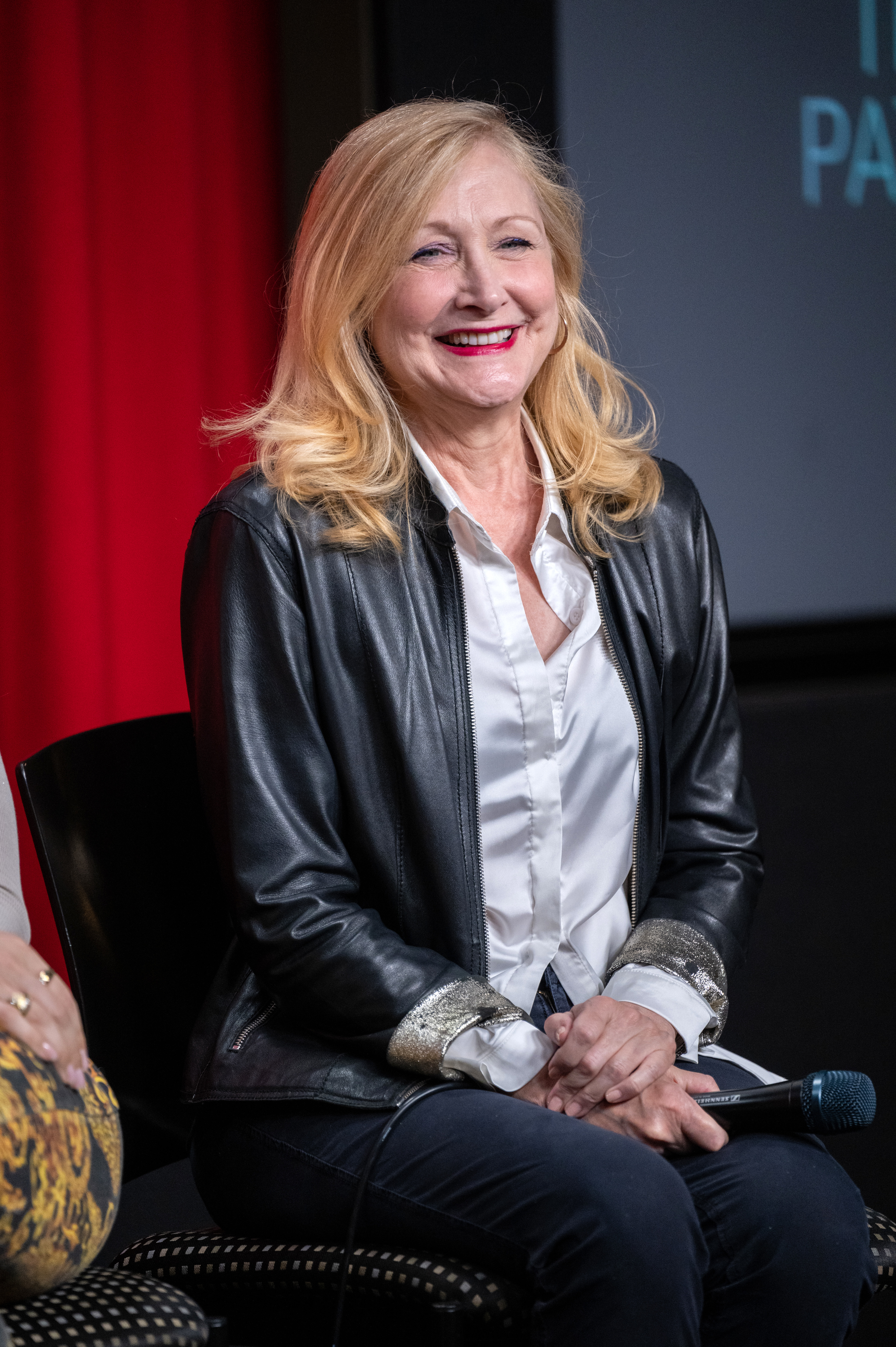 Woman seated, smiling, wearing a black leather jacket over a white blouse
