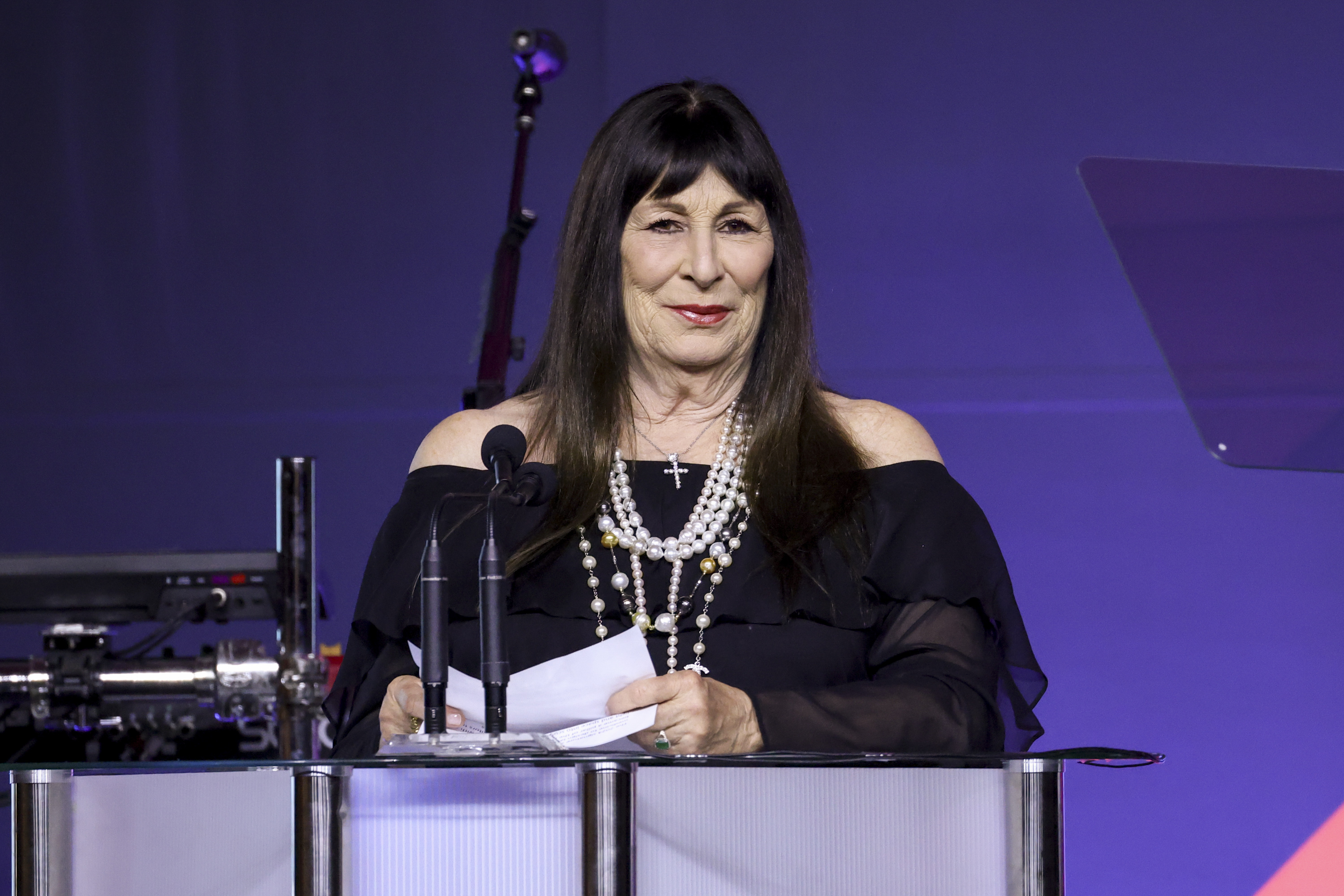 Anjelica Huston, wearing an off-shoulder dress with layered necklaces, stands at a podium speaking at an event