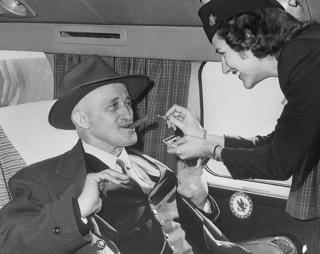 An airline stewardess lights a cigar for the passenger in a black hat and suit on an airplane
