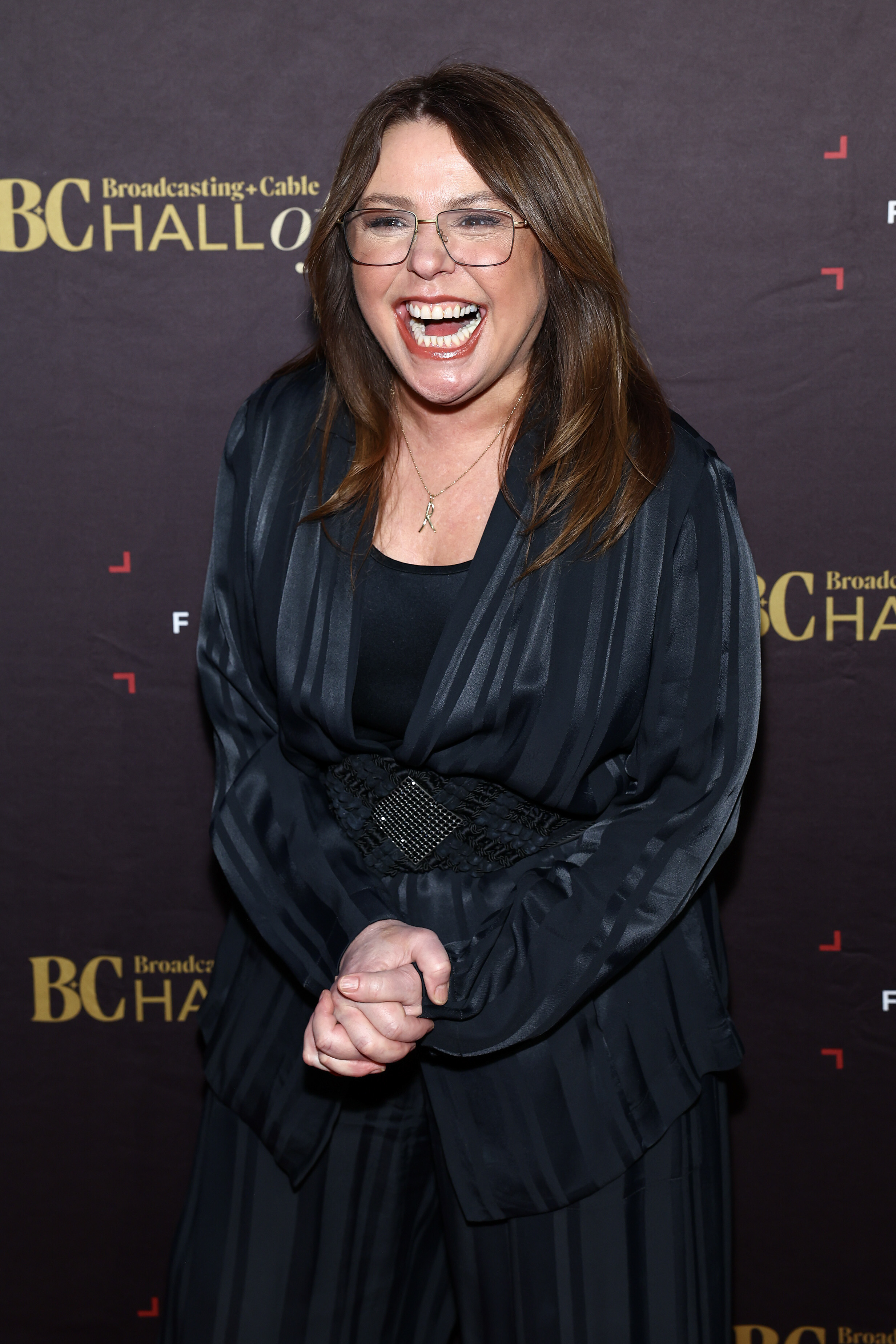 Rachael Ray stands on the red carpet at the Broadcasting & Cable Hall of Fame event, laughing, wearing a stylish, dark outfit with a belted waist