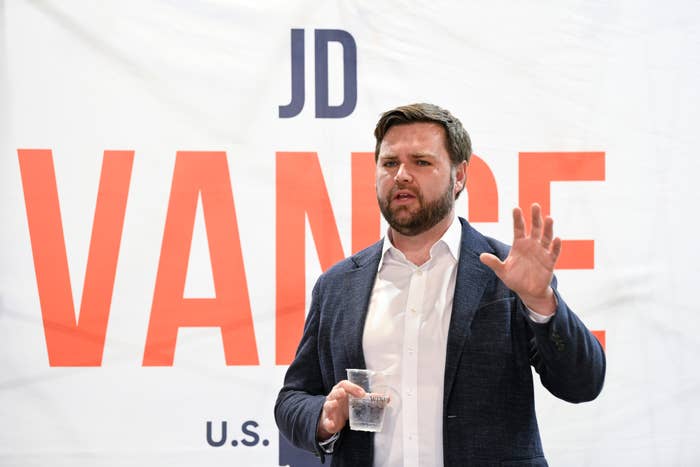 J.D. Vance speaking in front of a sign with his name, holding a clear plastic cup, wearing a white shirt and dark blazer