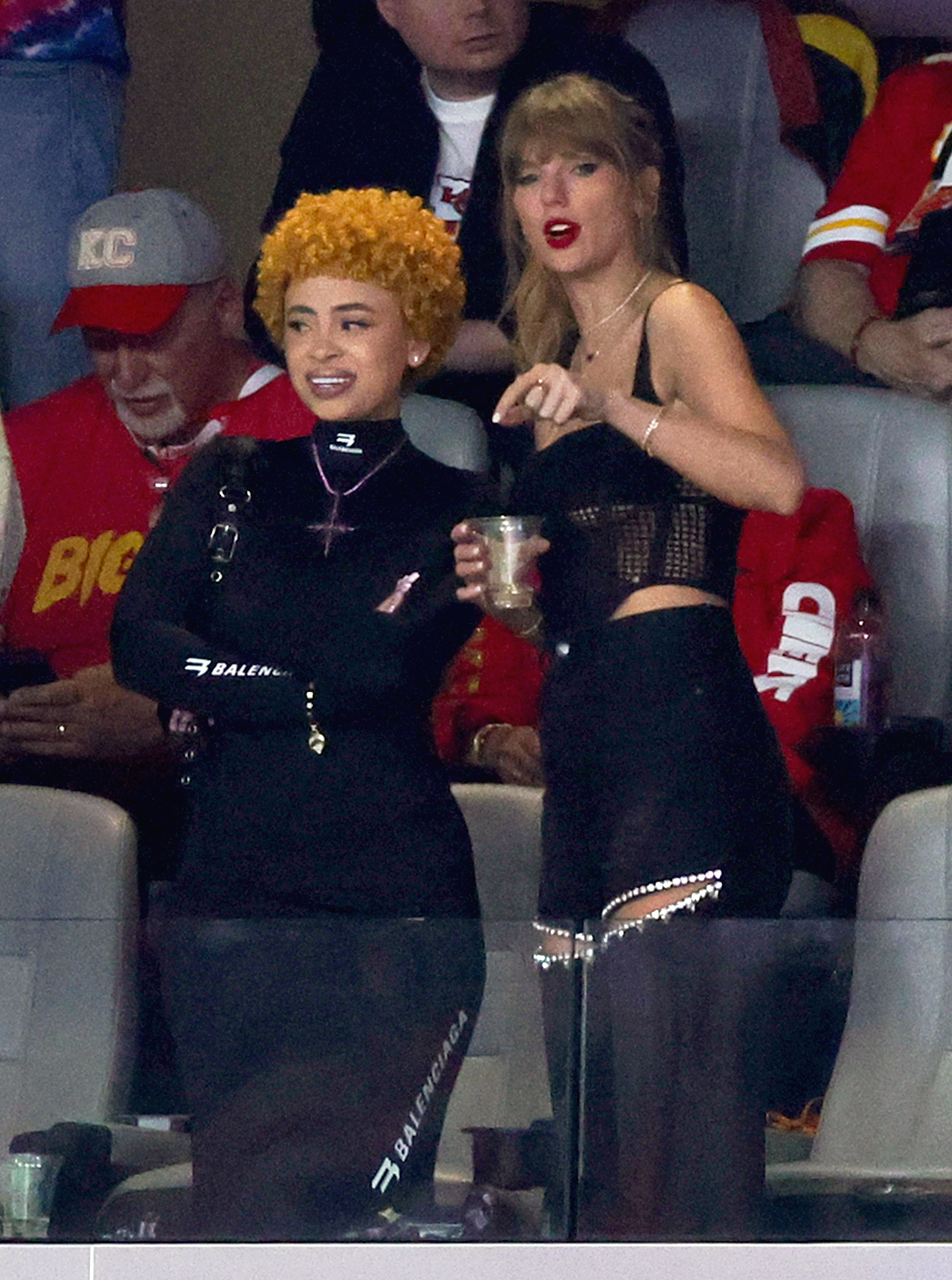 Ice Spice in a dark Balenciaga outfit and Taylor Swift in a dark mesh top with shorts, both standing and talking at a sports event