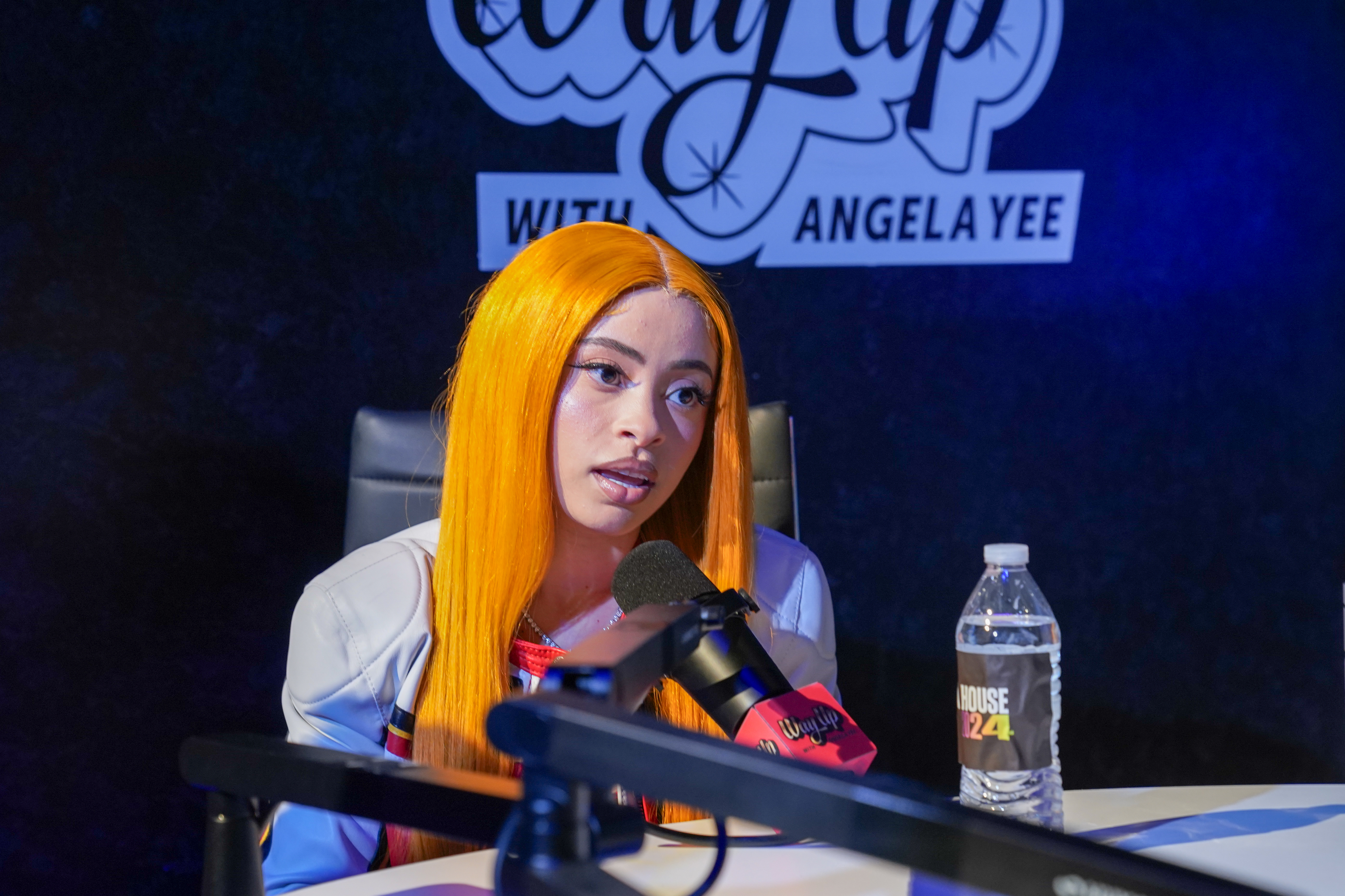 Ice Spice sits at a table speaking into a microphone during an interview with Angela Yee, with a water bottle in front of her
