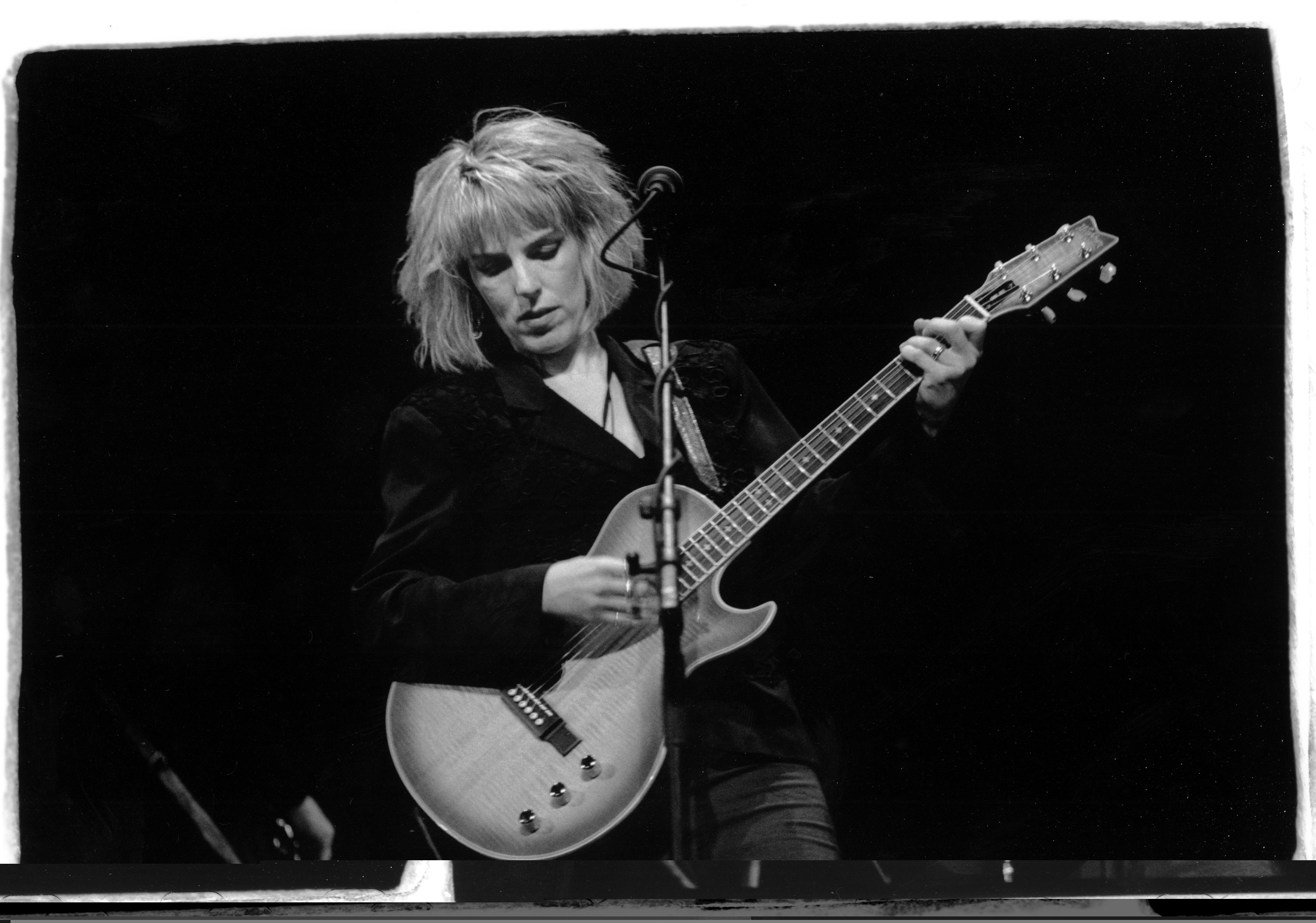 Lucinda Williams playing a guitar on stage, focused and expressive