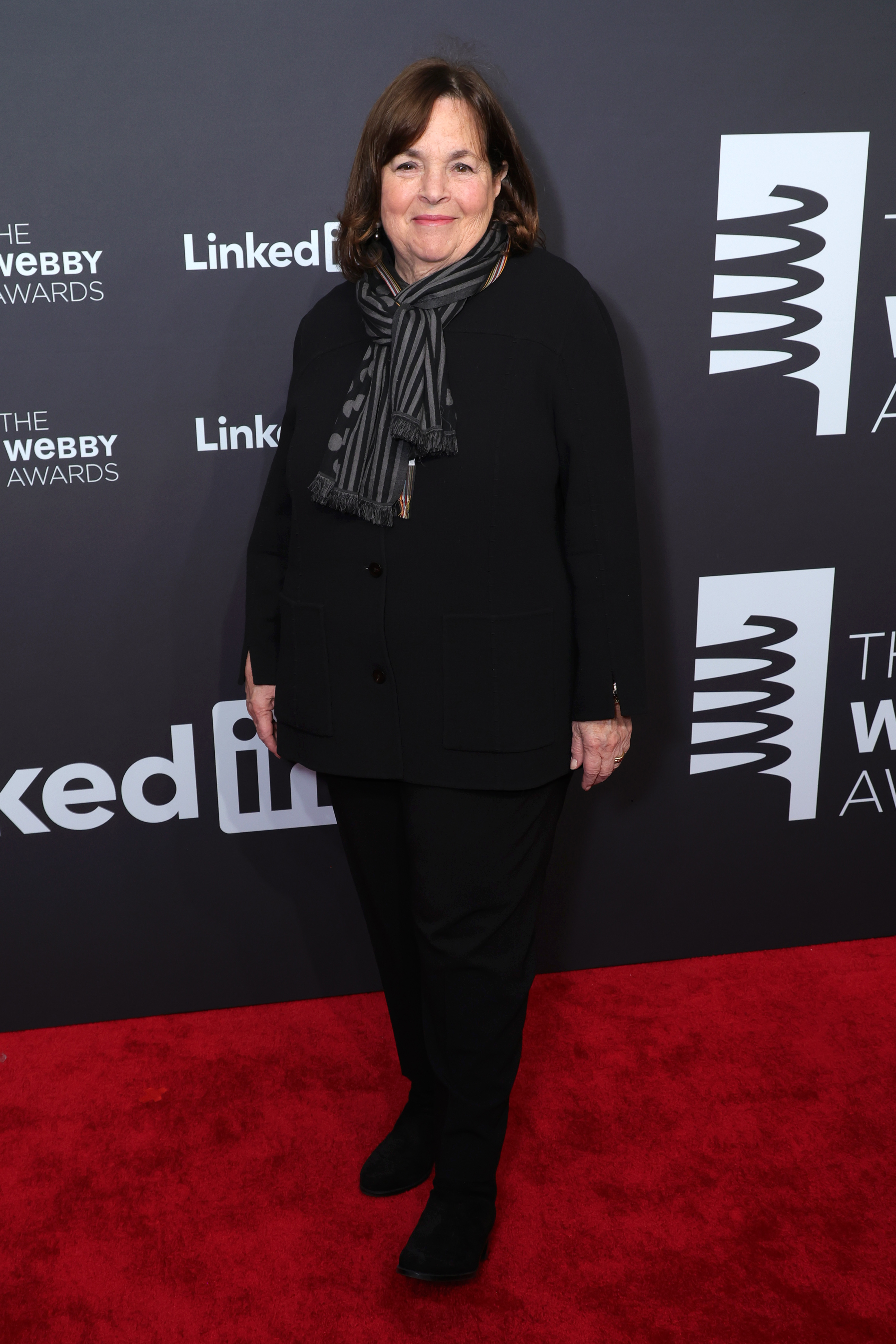 Ina Garten poses on the red carpet at The Webby Awards in a black outfit with a striped scarf