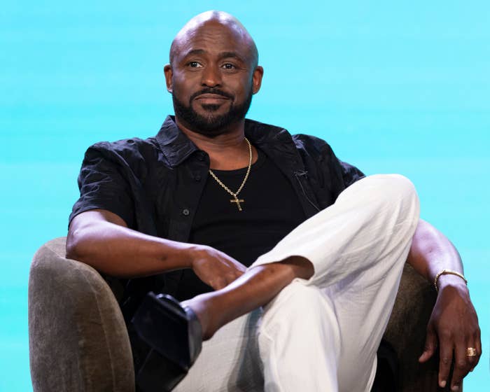 Wayne Brady sits cross-legged on a stage chair, wearing a dark shirt, light pants, a cross necklace, and a relaxed expression
