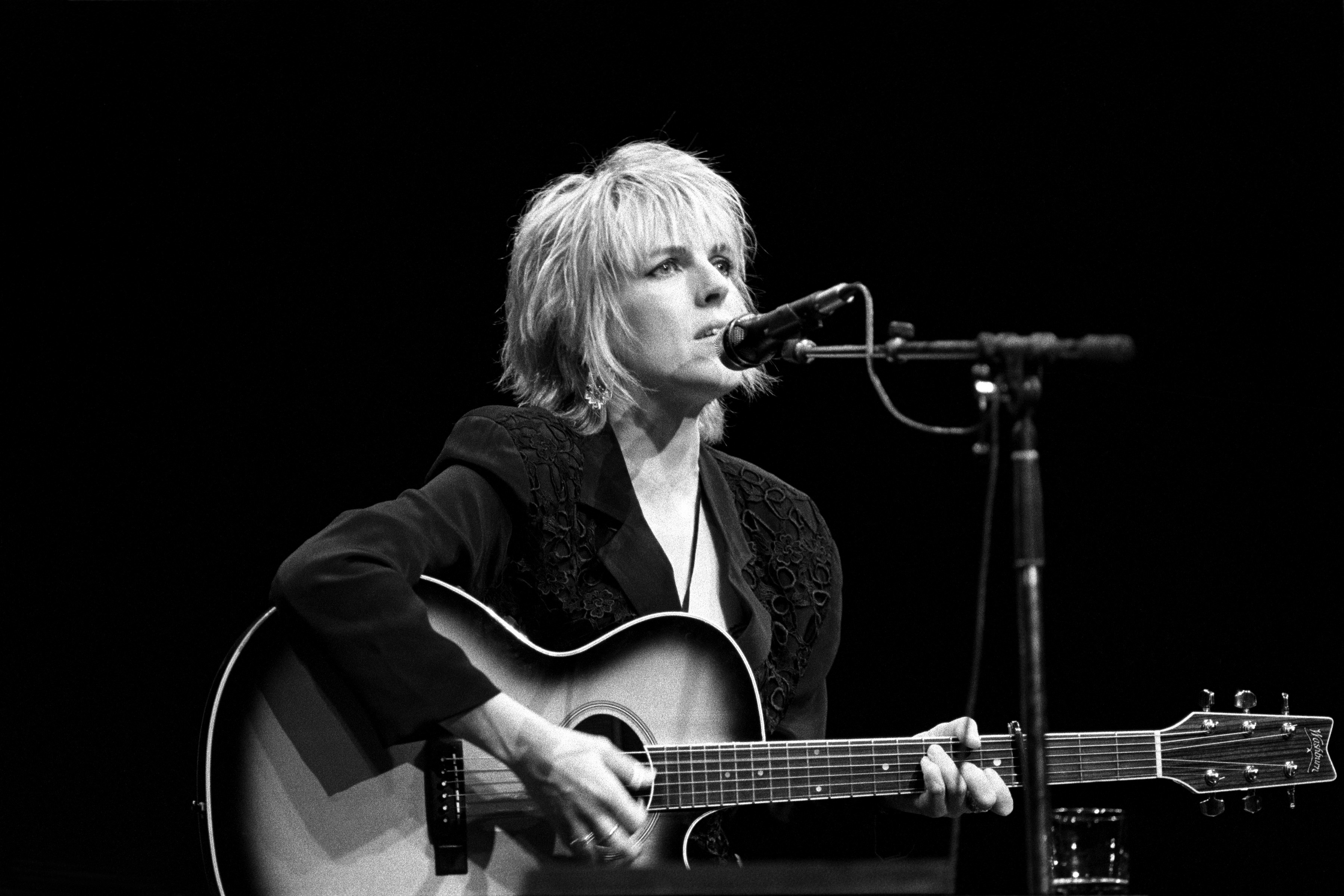 Lucinda Williams plays an acoustic guitar and sings into a microphone during a live performance