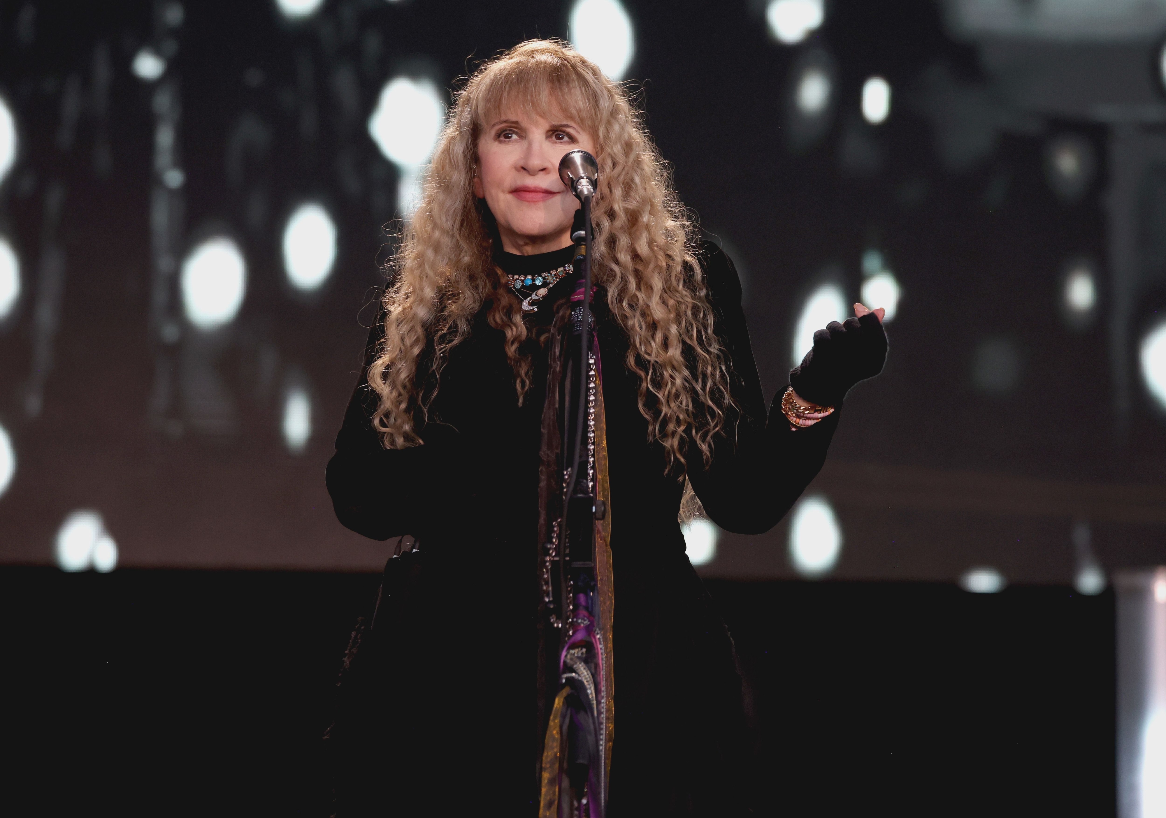 Stevie Nicks stands on stage, wearing a black outfit with a scarf, performing with a microphone