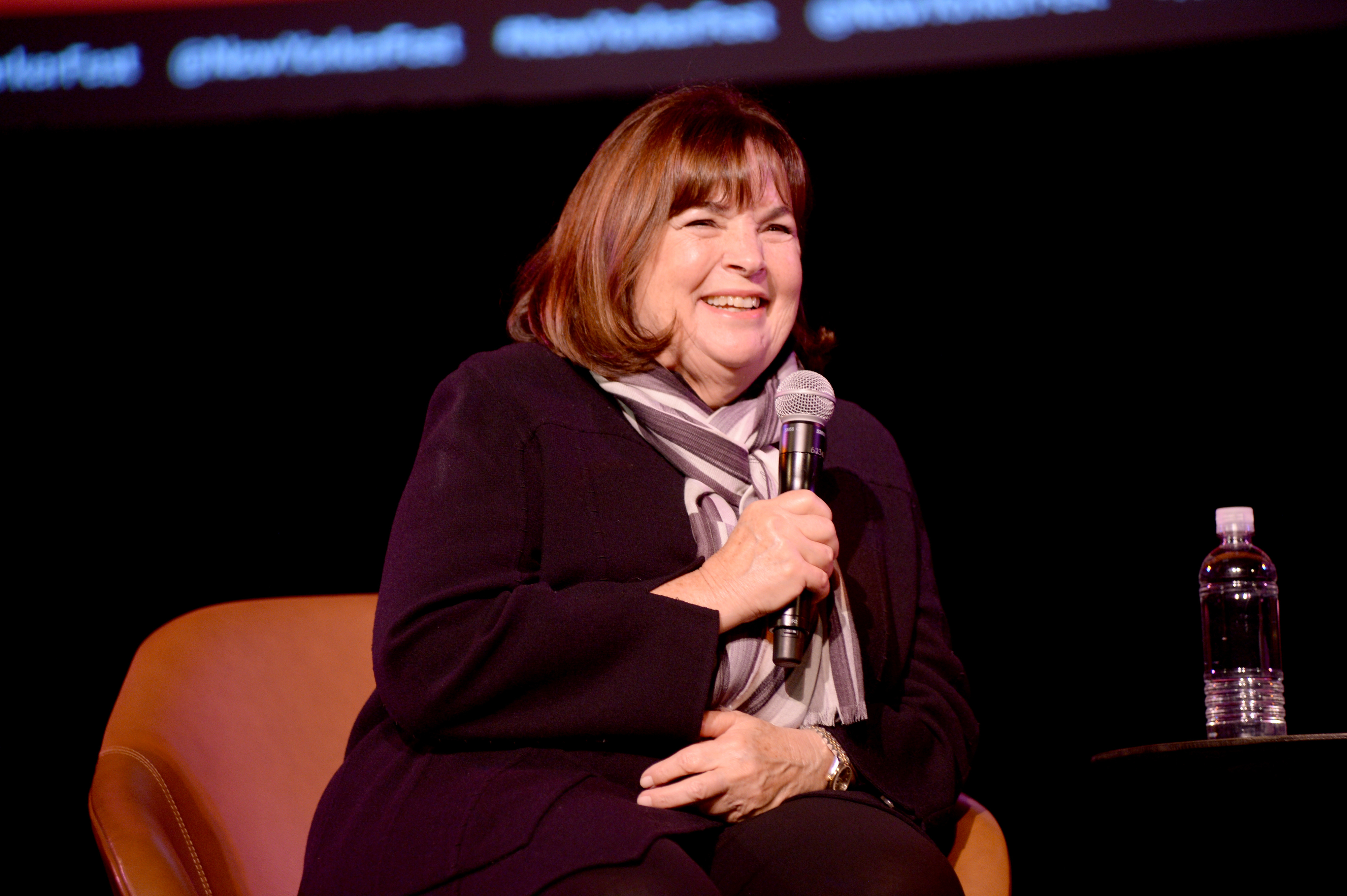 Ina Garten, seated and smiling, holds a microphone during a speaking event. She is dressed in a dark outfit and a striped scarf. A water bottle is on the table beside her