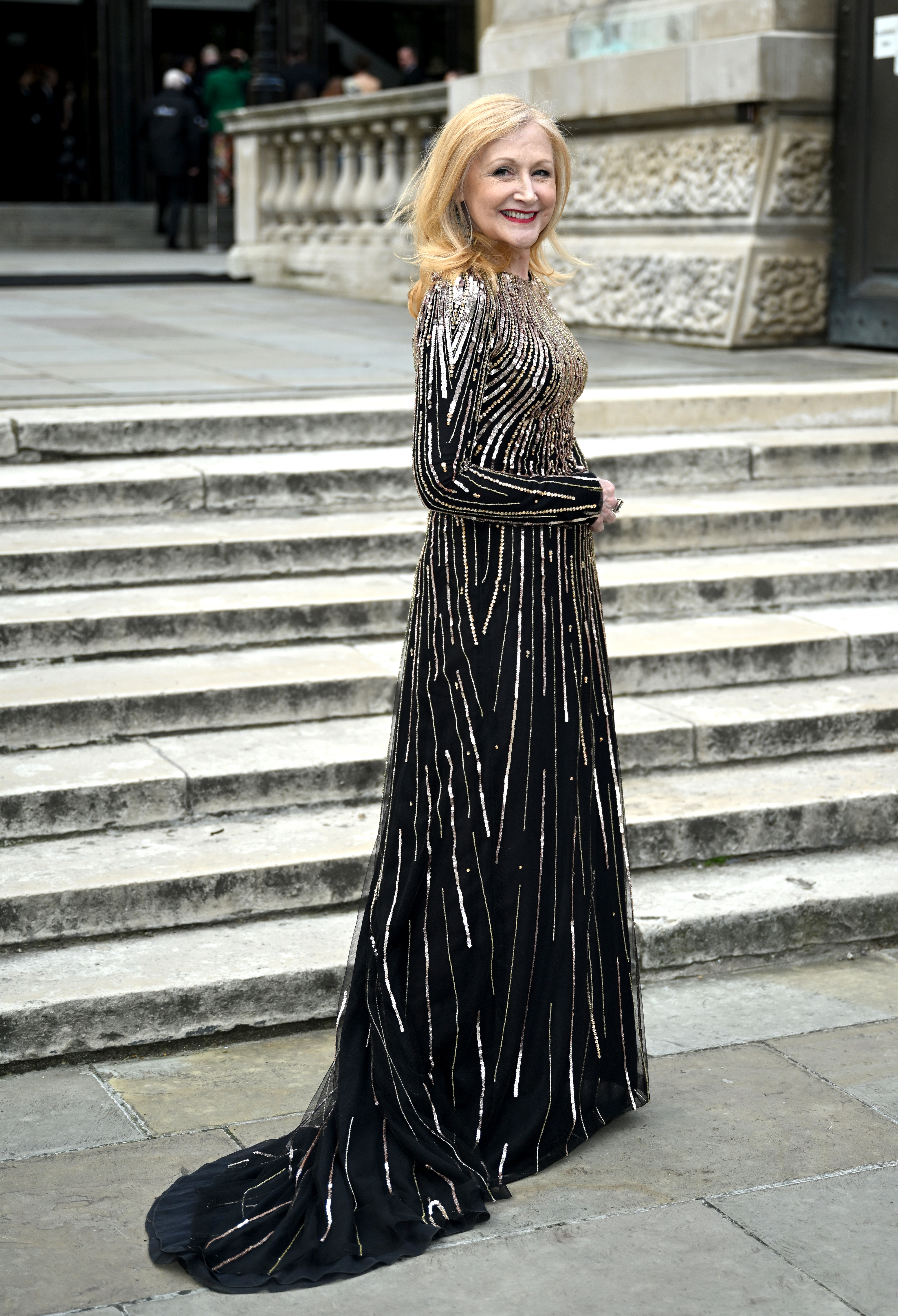 A woman in a long, elegant gown with metallic vertical stripes poses on a stone staircase
