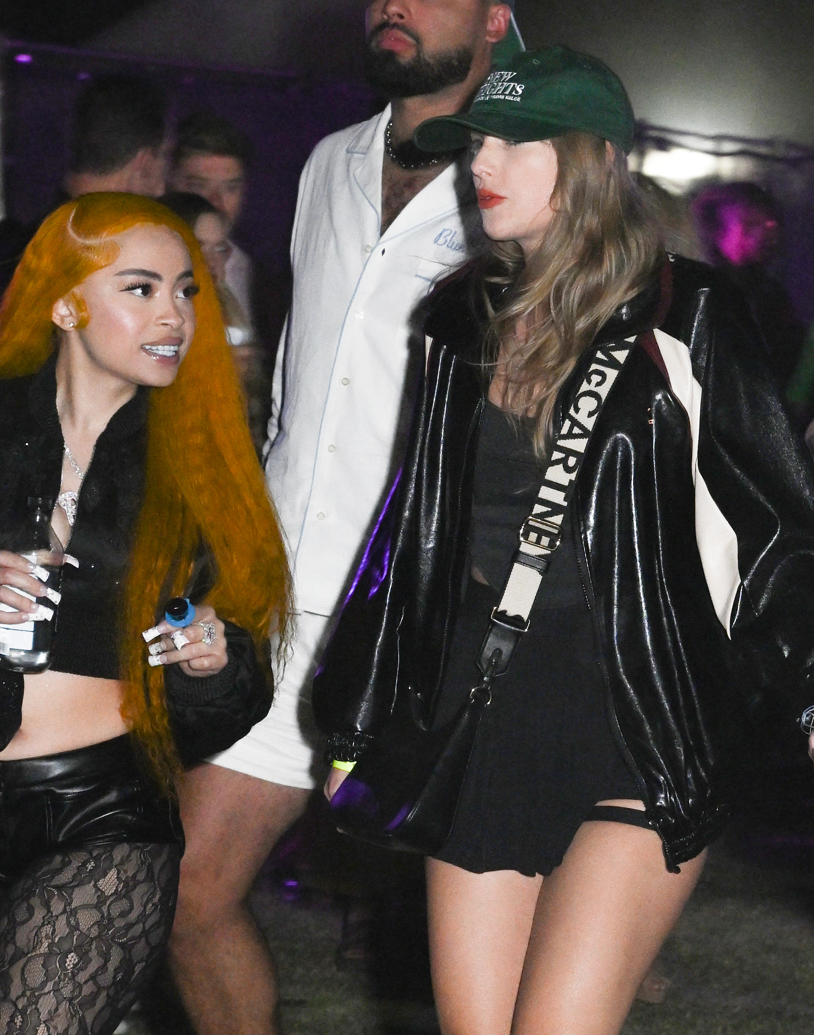 Ice Spice wearing a dark crop top and lace pants, and Taylor Swift wearing a black dress with a white Stella McCartney bag, seen together at an event