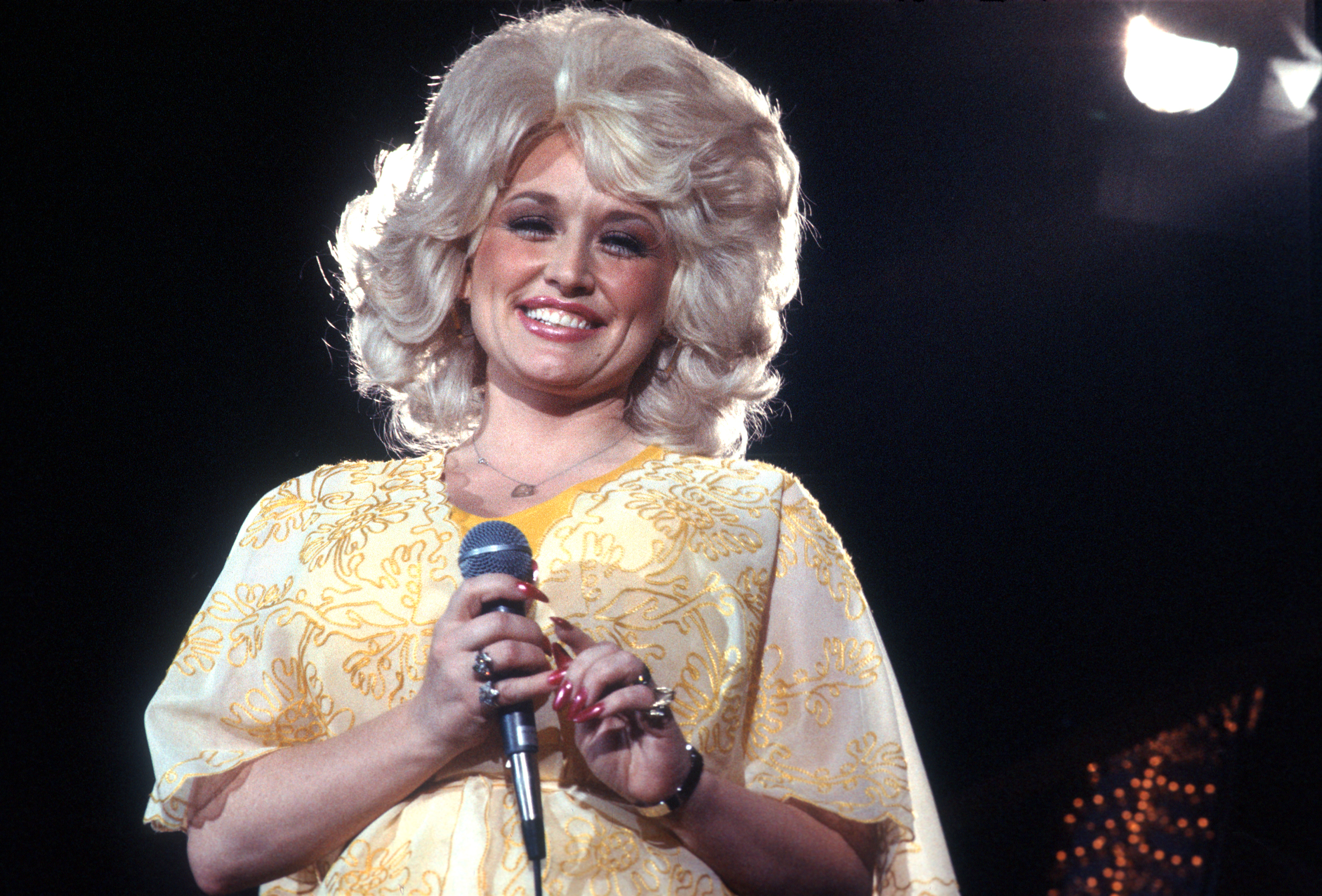 Dolly Parton smiles and holds a microphone, wearing a patterned dress with voluminous hair, under stage lights