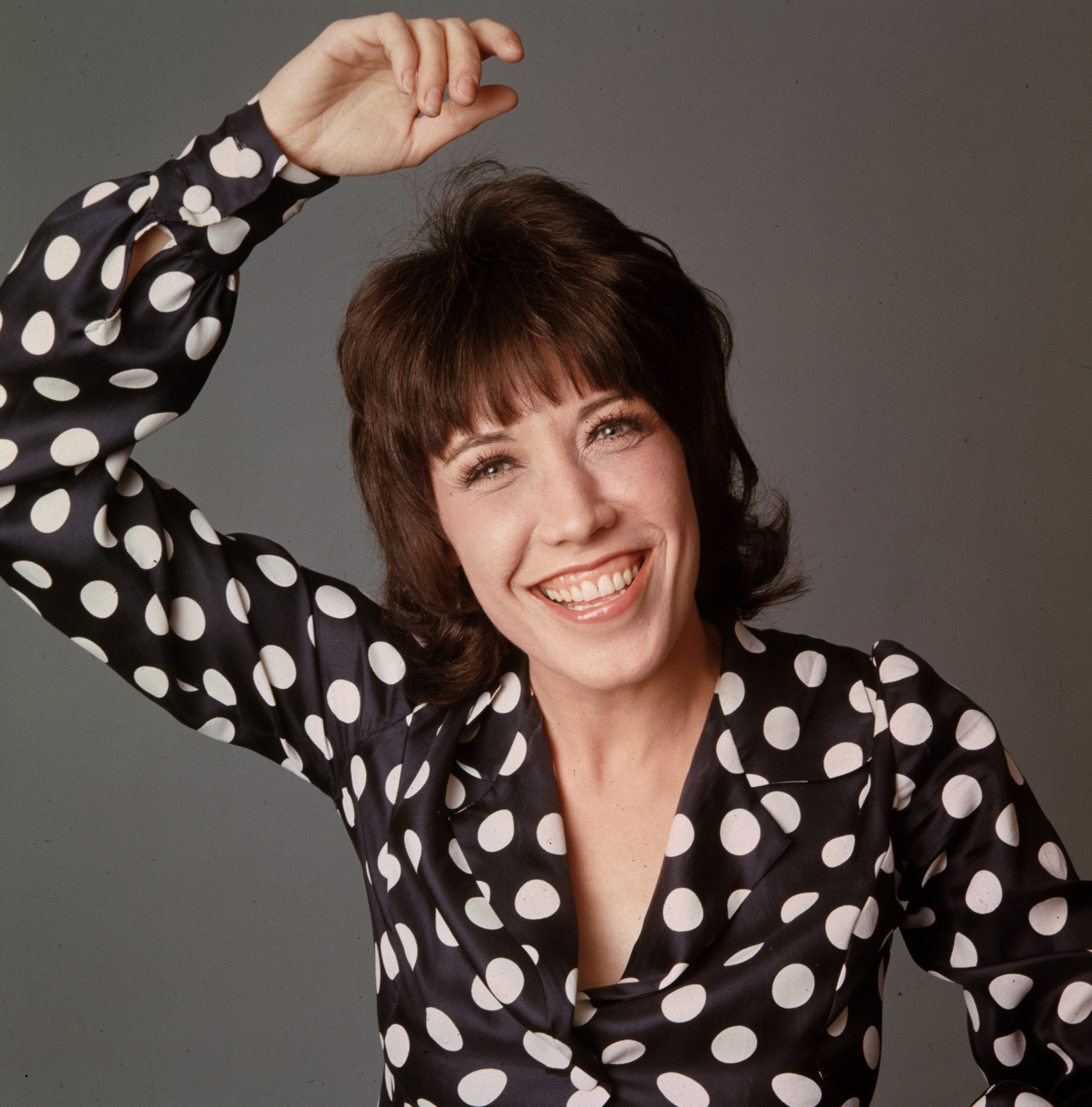 A woman with short brown hair smiles at the camera. She is wearing a polka dot blouse and raising one arm