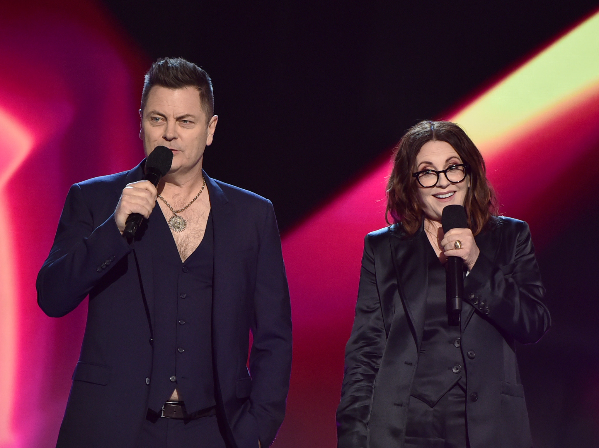 Nick Offerman and Megan Mullally perform on stage, both dressed in dark suits and holding microphones