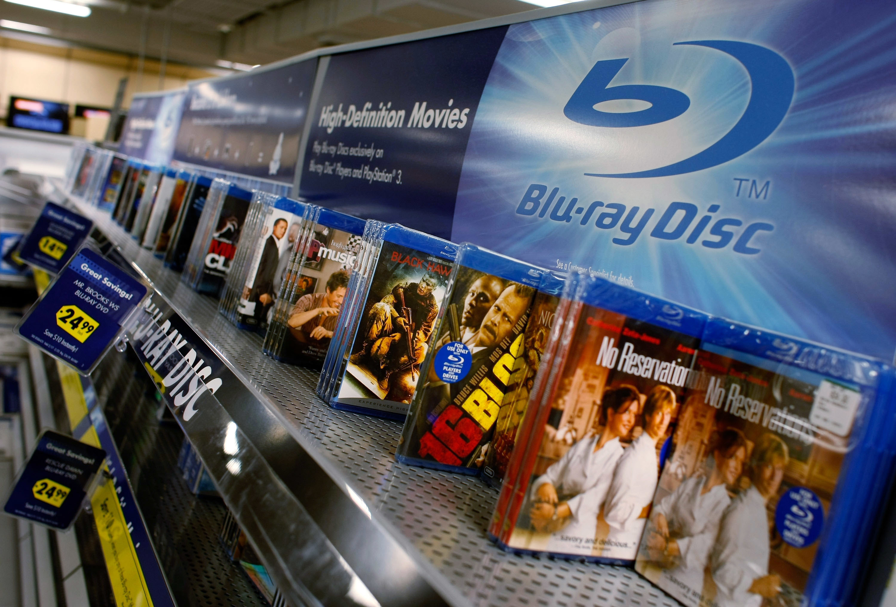 A display shelf in a store holds various Blu-ray movie discs, including "The Big Sick" and "No Reservations," under a sign that reads "High-Definition Movies."