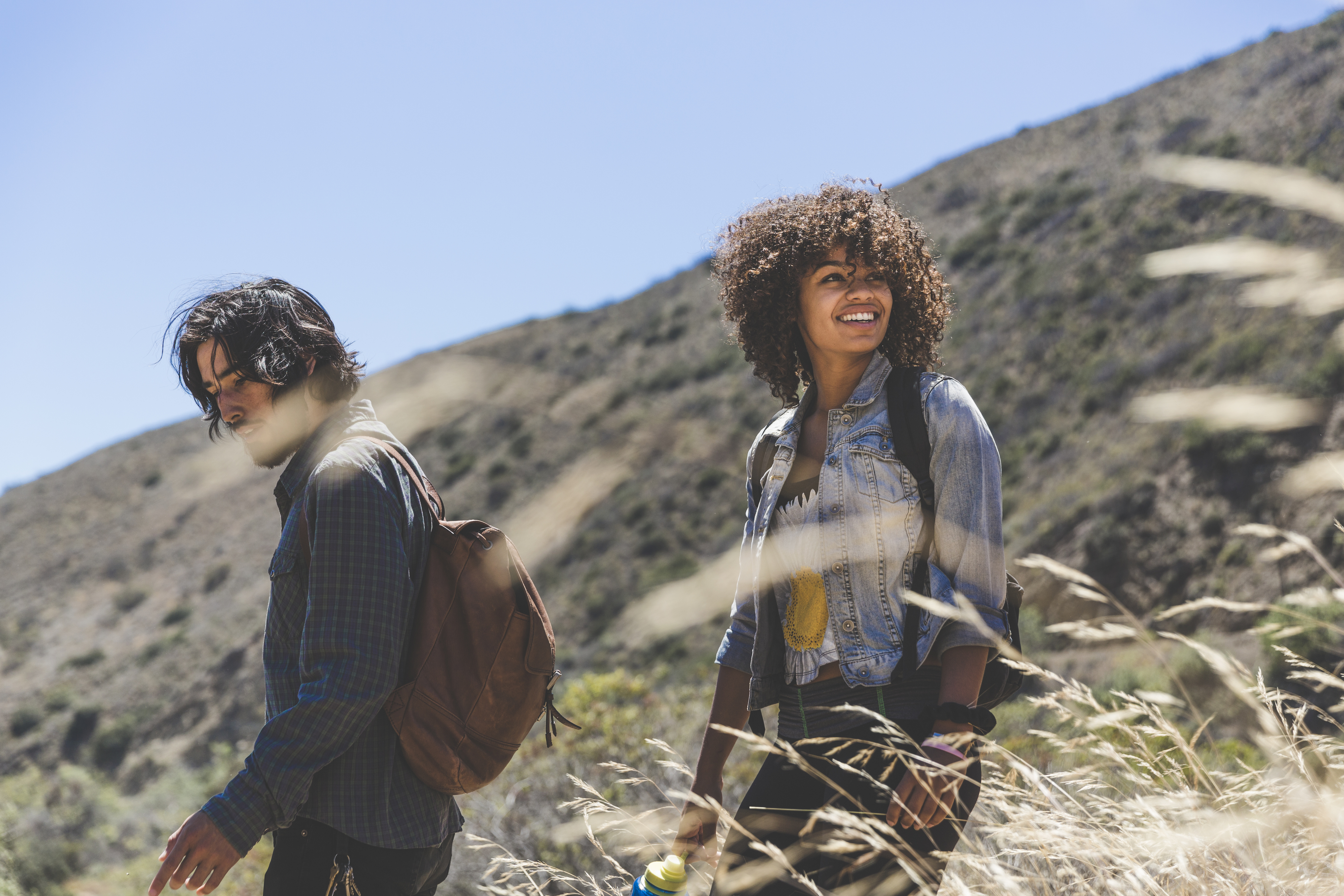 A couple walk through a grassy field with smiles. They are casually dressed and carrying backpacks