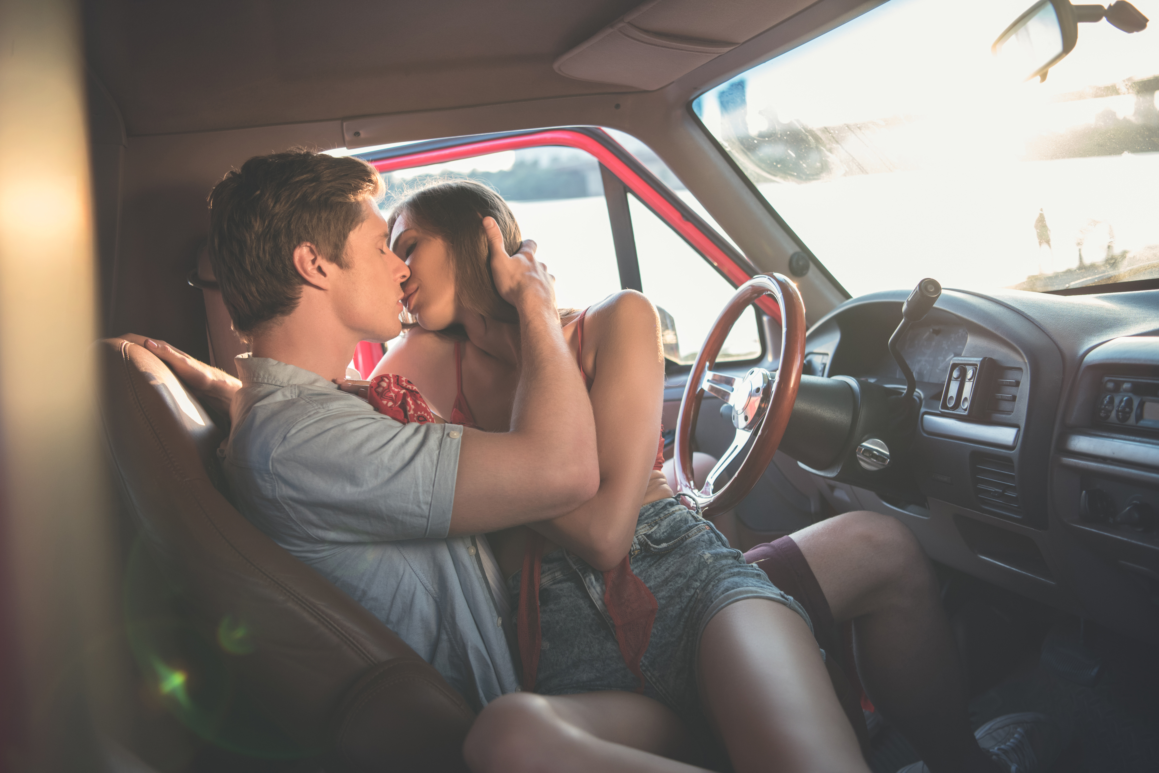 A young couple is passionately kissing inside a truck, with the man's hand gently holding the side of the woman's face. Sunlight streams through the window