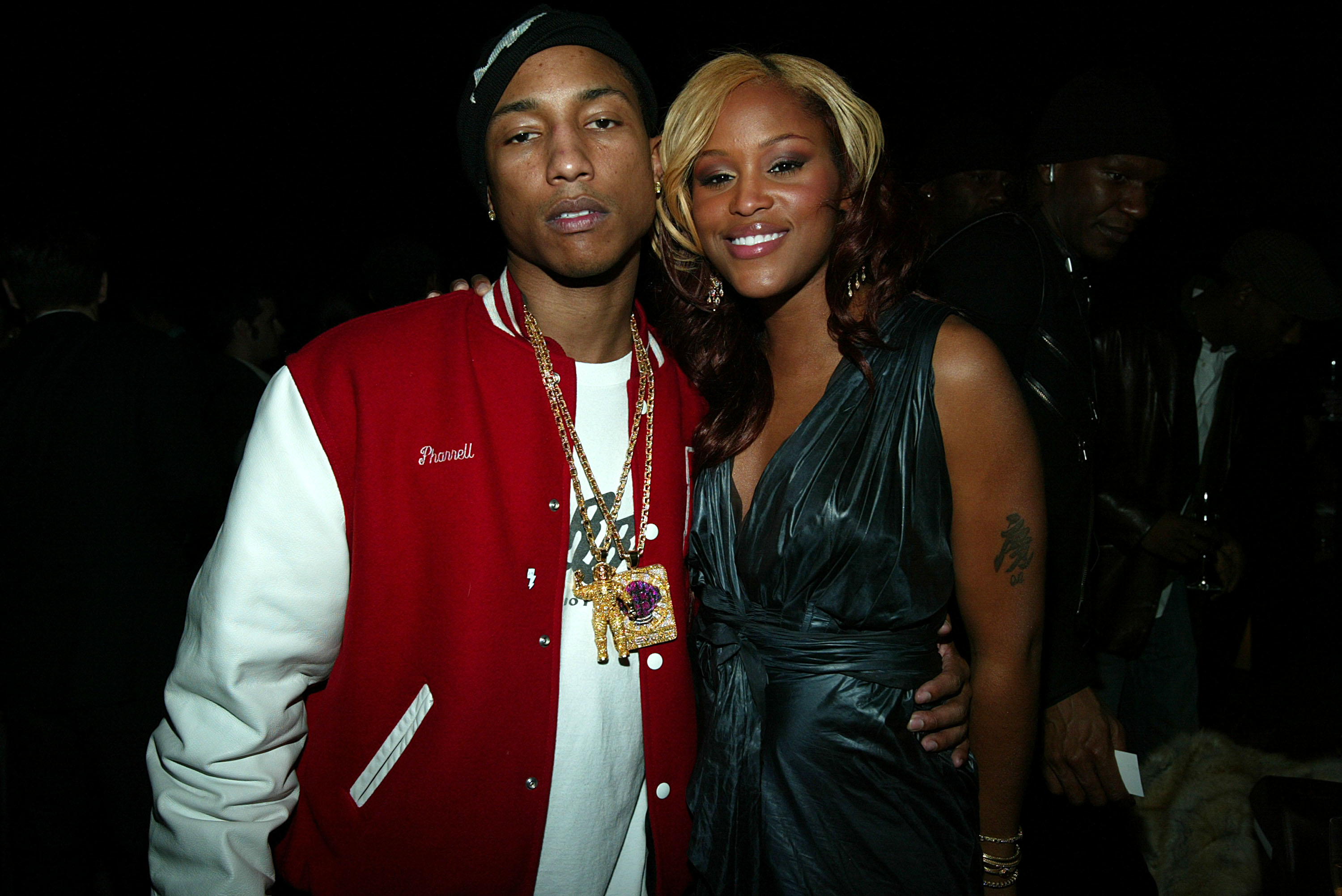 Pharrell Williams in a red jacket and Eve in a sleeveless dress at a social event, posing together and smiling