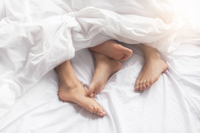 Two pairs of legs intertwined under white sheets, suggesting intimacy in a bedroom setting. Faces are not visible