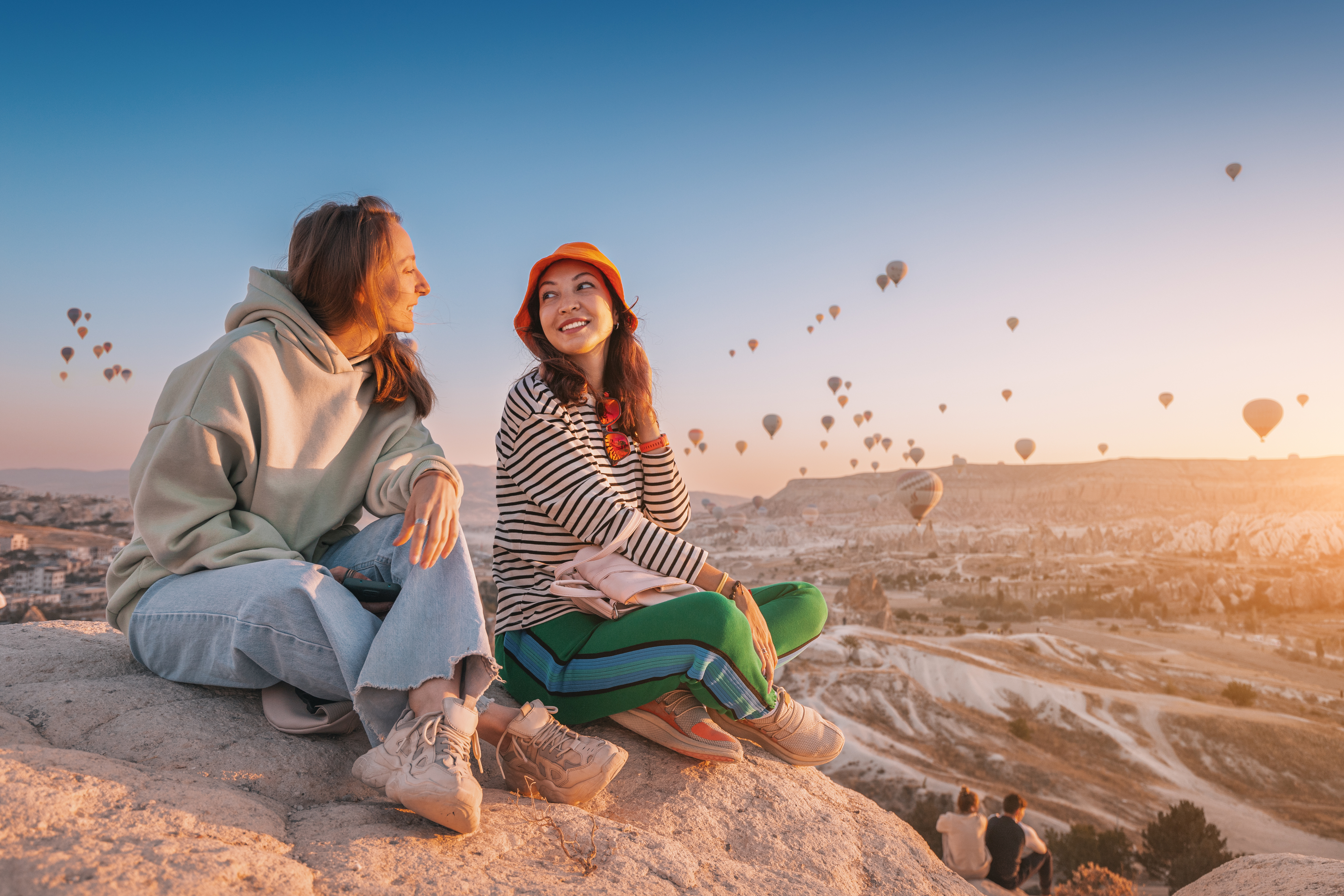 Two women smiling at each other, sitting on a rock with hot air balloons in the background at sunrise