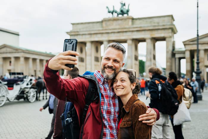A man and woman smile while taking a selfie in front of the Brandenburg Gate in Berlin, Germany