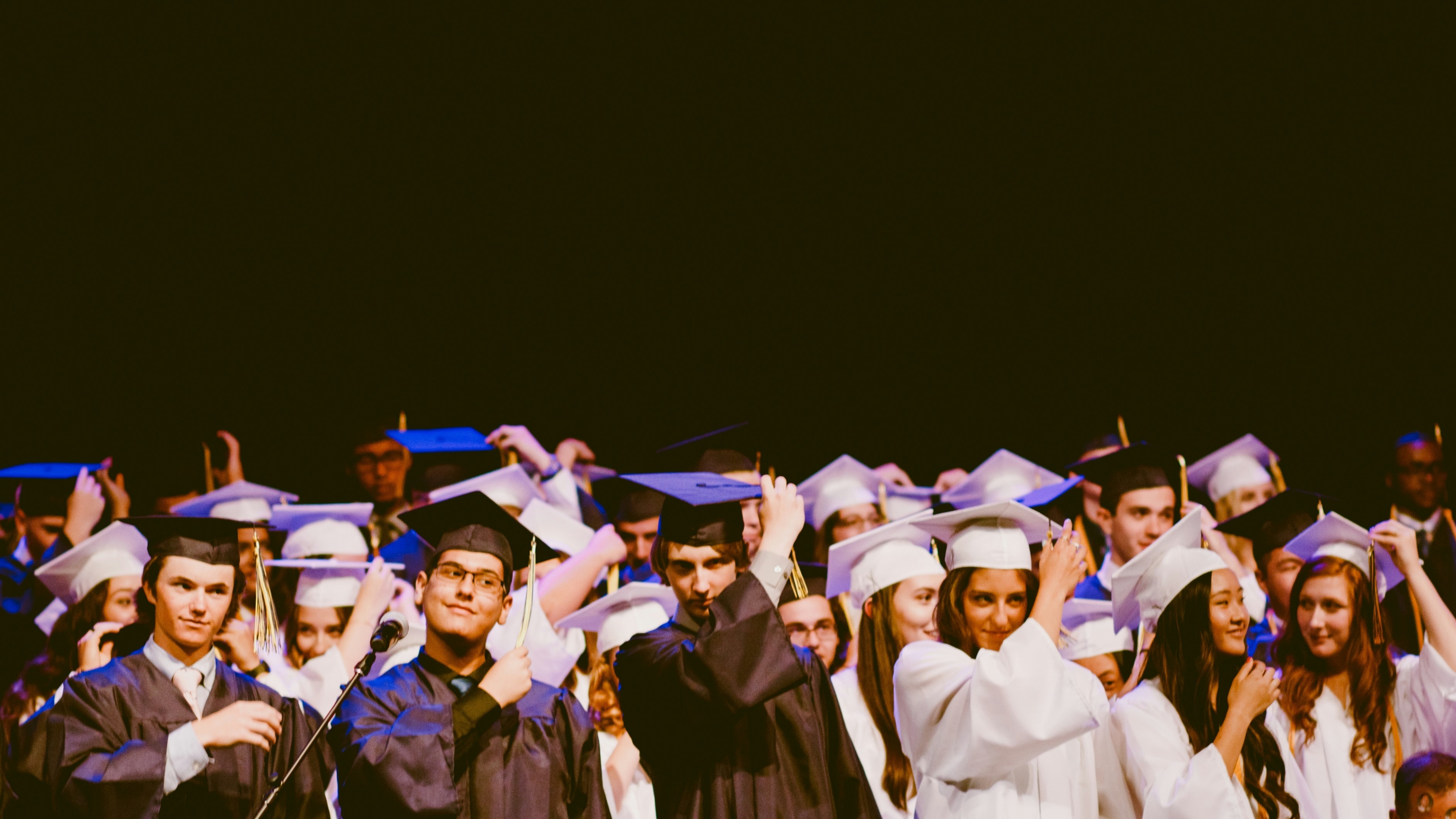 A large group of graduates in caps and gowns celebrates on stage, adjusting their tassels