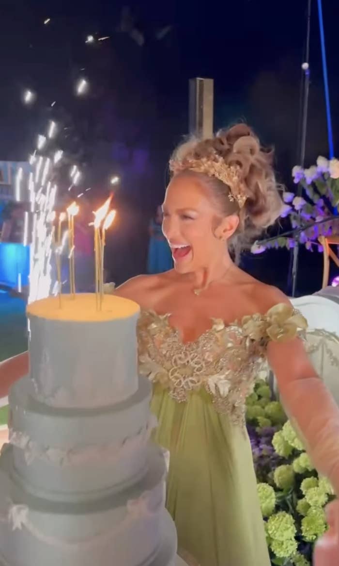 Jennifer Lopez in ornate, light gown stands behind a multi-tiered cake with lit candles and sparklers, smiling joyfully