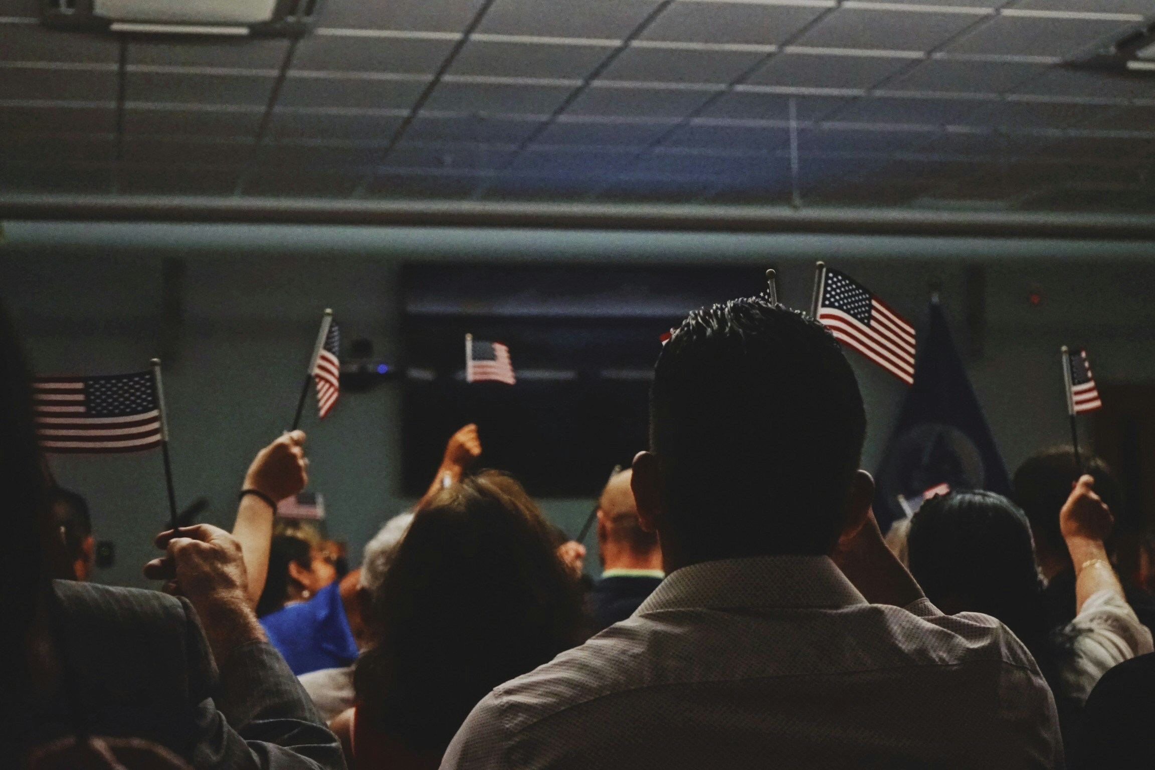 A group of people in a room hold small American flags, raising them in unison in a naturalization ceremony