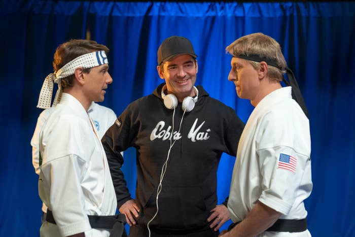 Ralph Macchio (left) and William Zabka (right) in karate outfits with headbands standing with a man wearing a "Cobra Kai" hoodie and headphones around his neck