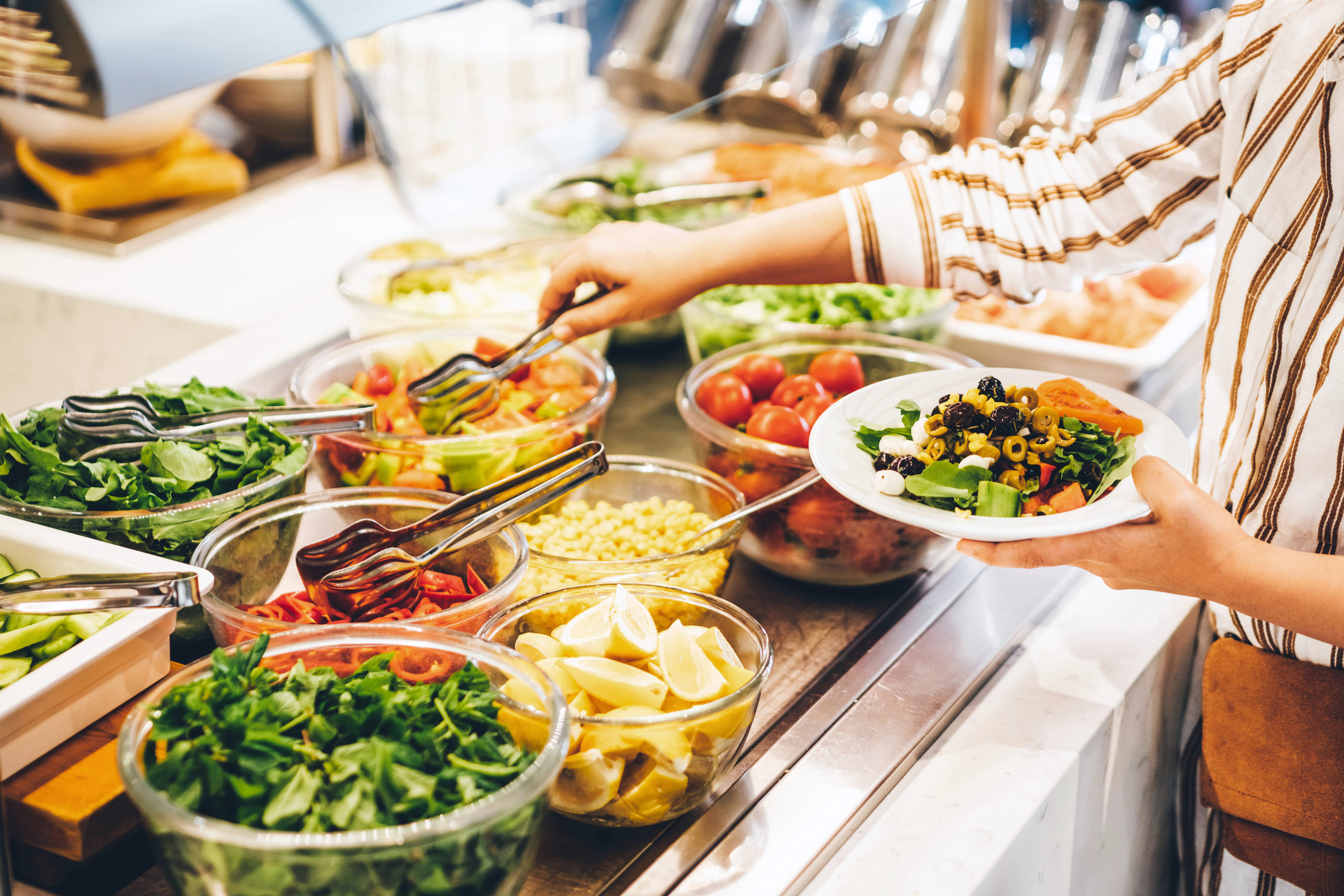 Person serving themselves from a buffet with various fresh vegetables, greens, corn, and lemon wedges into a white plate
