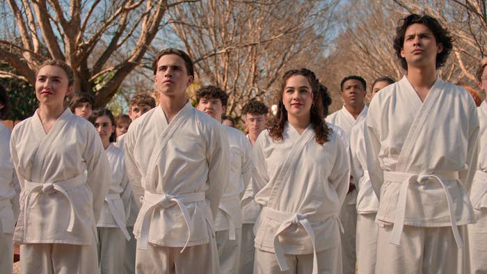 Multiple characters from "Cobra Kai" in white karate gis stand in formation outside, focused and determined