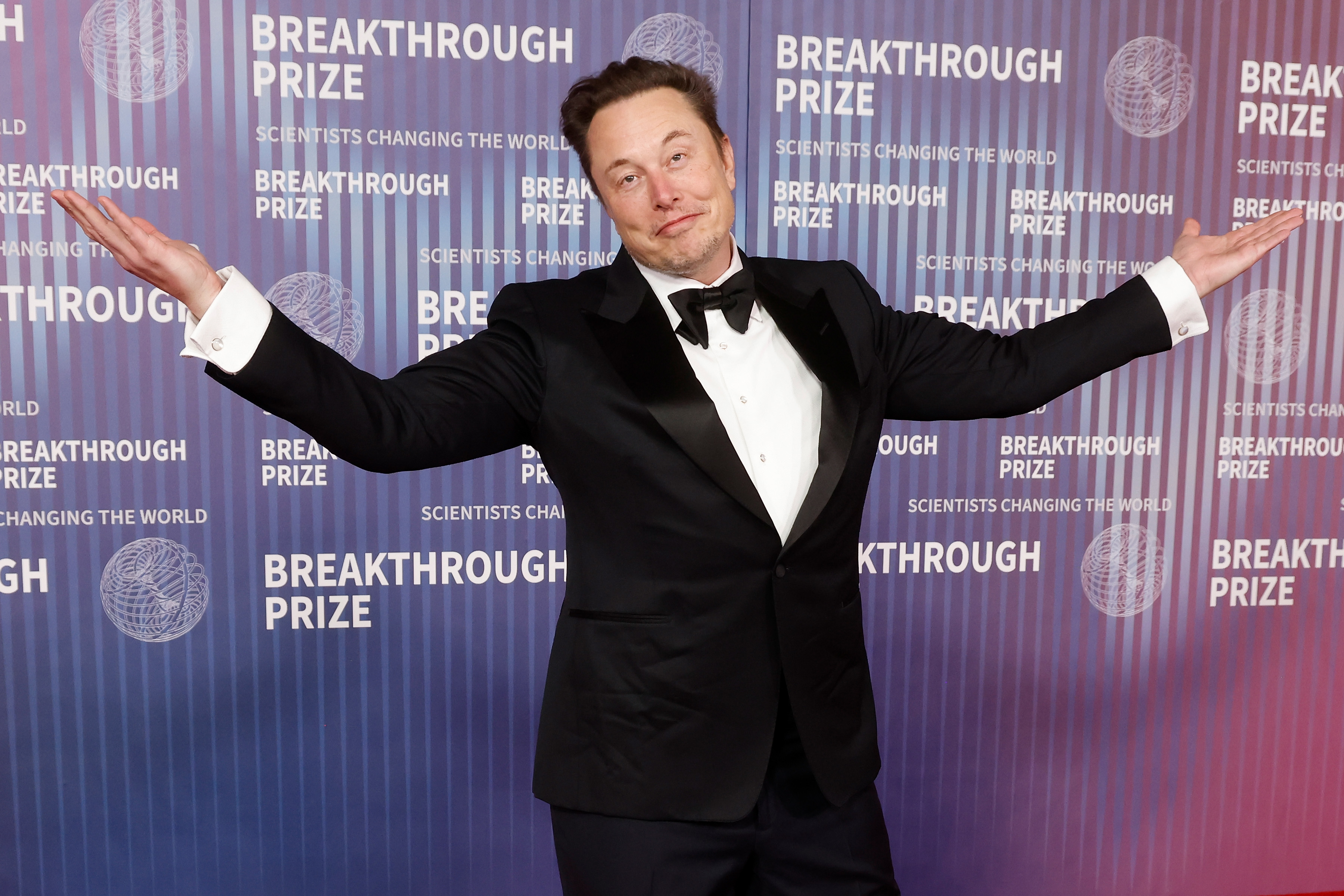 Elon Musk on the red carpet at the Breakthrough Prize event, wearing a black tuxedo with a white dress shirt and black bow tie, gesturing with arms wide open