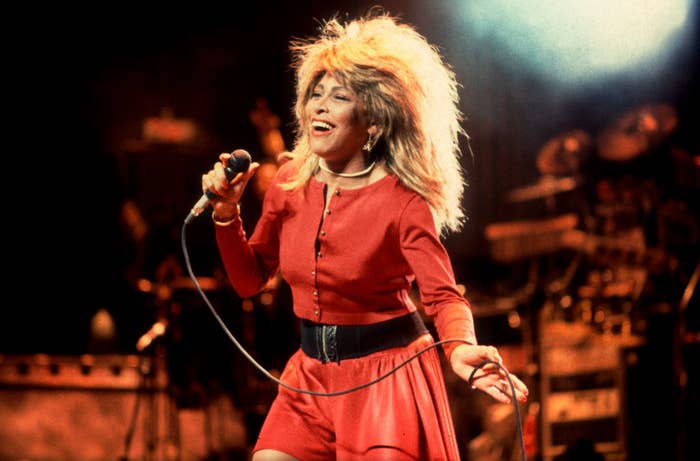 Tina Turner performing on stage, wearing a stylish red outfit with a wide black belt, holding a microphone and singing passionately