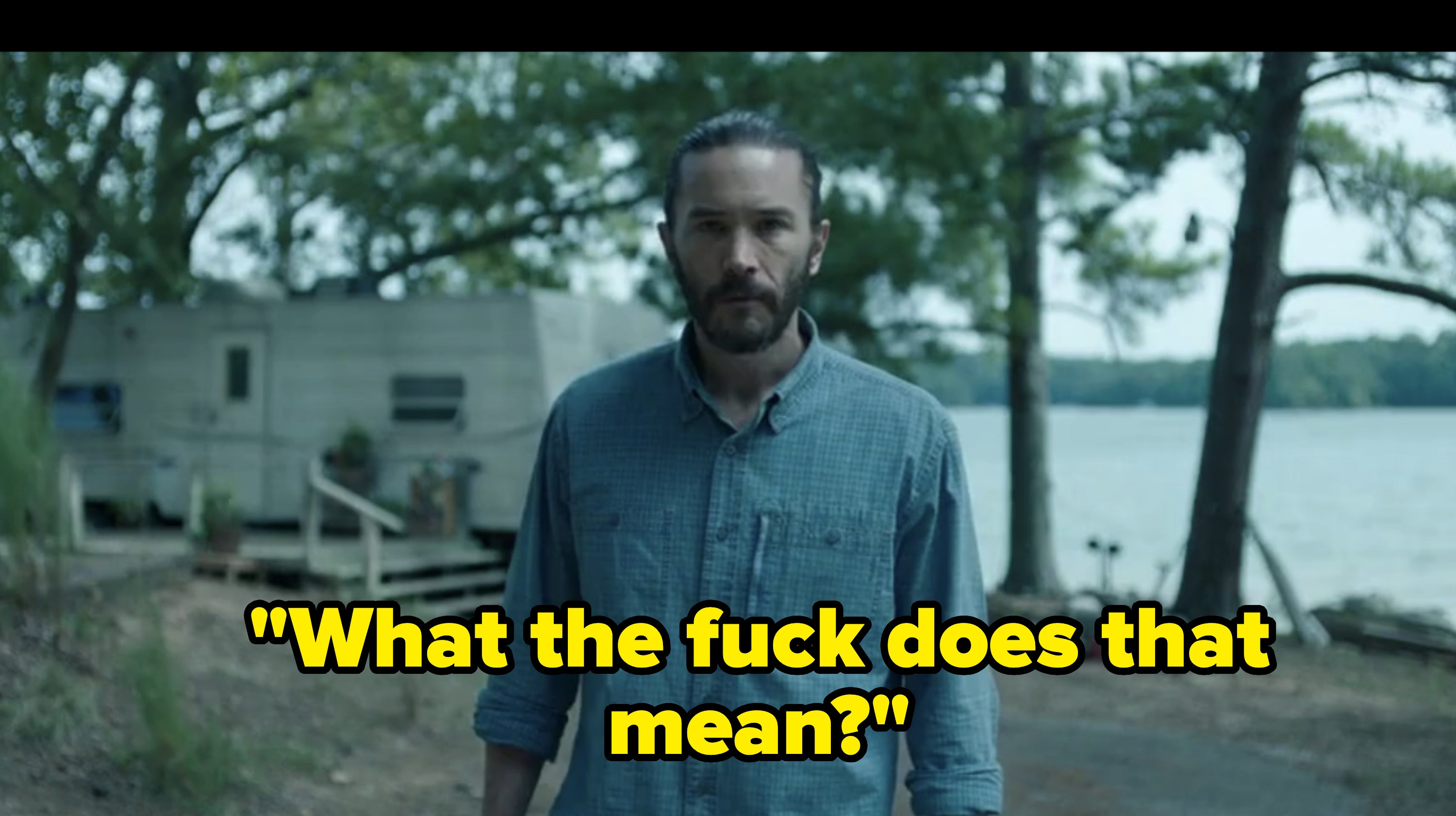 A man with a beard and ponytail, wearing a button-up shirt, stands outside near a lake and a trailer. Trees and water are visible in the background