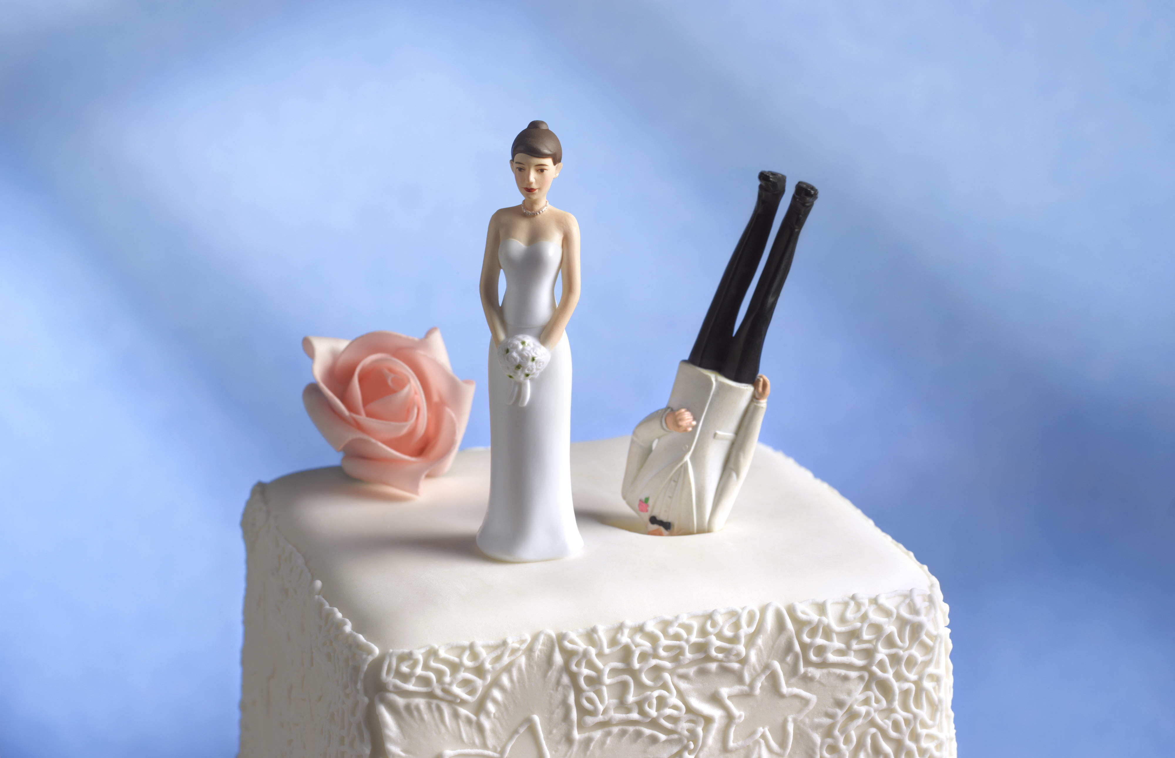 Bride and groom cake toppers on a wedding cake. The groom is upside down with his legs in the air while the bride stands holding a bouquet. A rose decoration is nearby