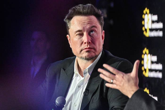 Elon Musk making a winking expression while holding a microphone during a speaking engagement