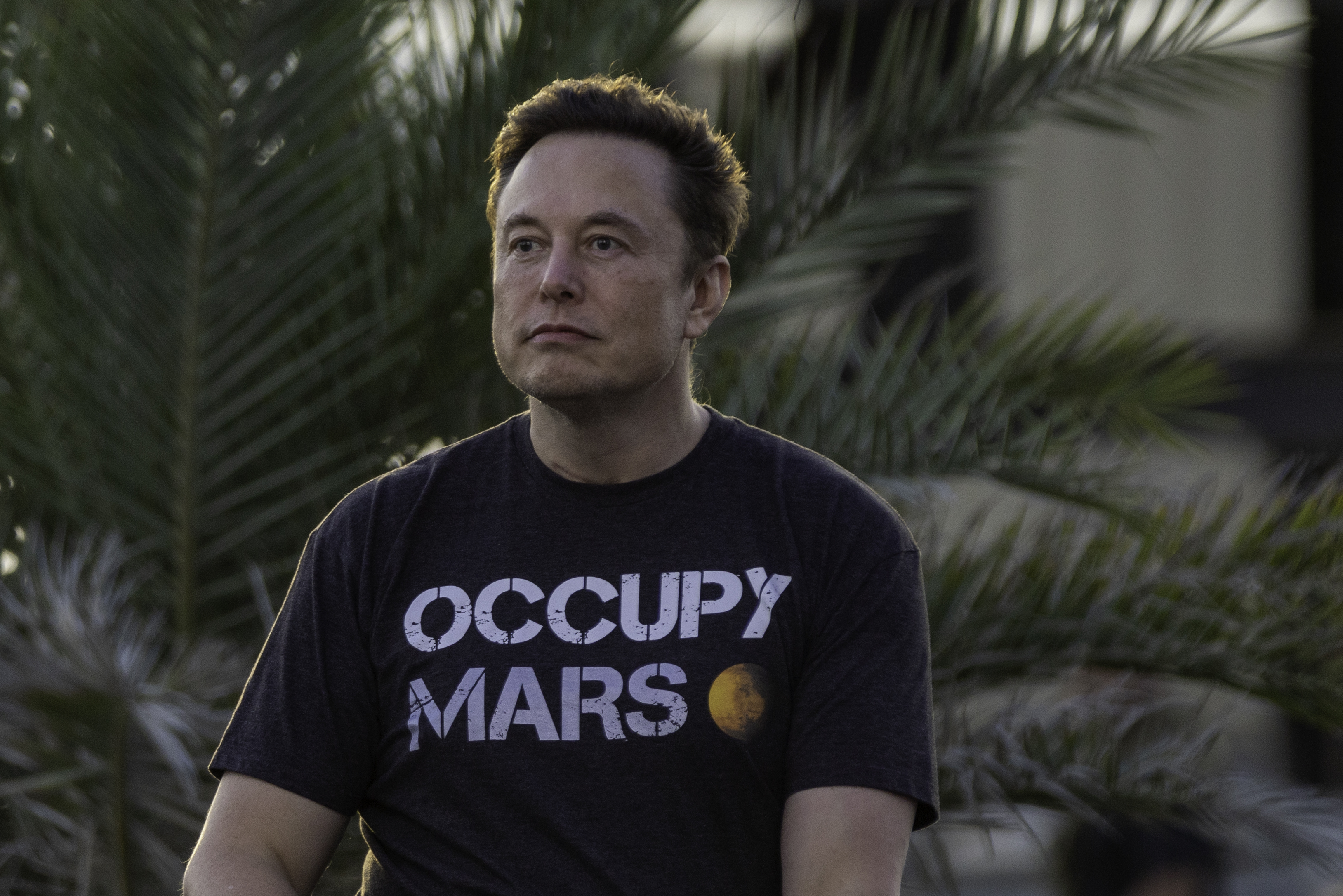 Elon Musk wearing an "Occupy Mars" T-shirt, outdoors with palm trees in the background