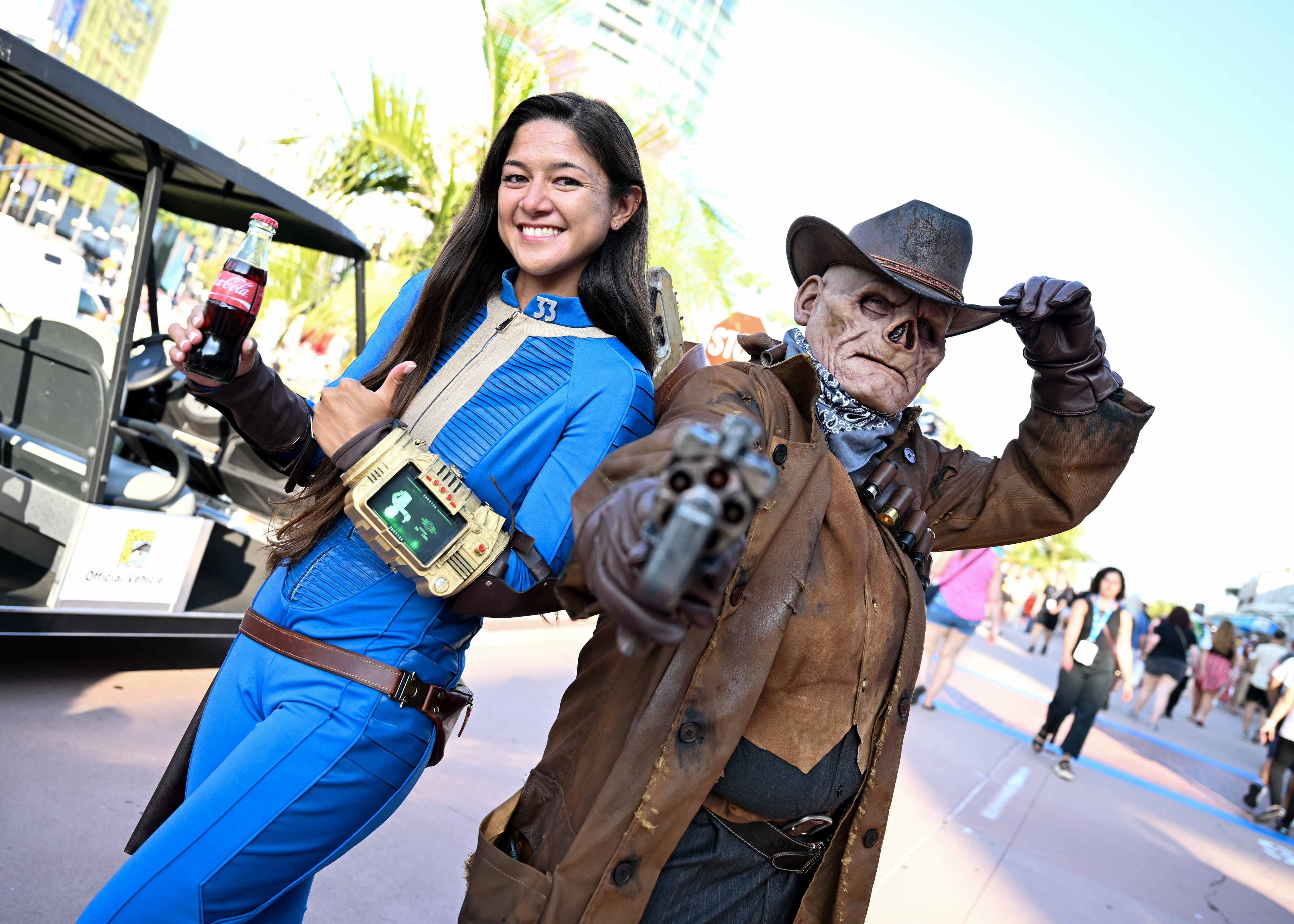Person dressed as a Vault Dweller from Fallout stands next to a person in a cowboy costume holding a gun and tipping their hat, both posing for a photo outside