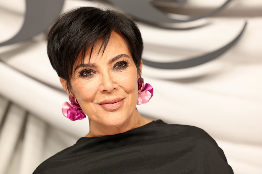 Kris Jenner smiles wearing a black top and large pink flower earrings